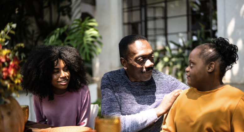 A father sits outdoors with his two adult daughters, resting a supportive hand on one daughter's shoulder as she speaks.