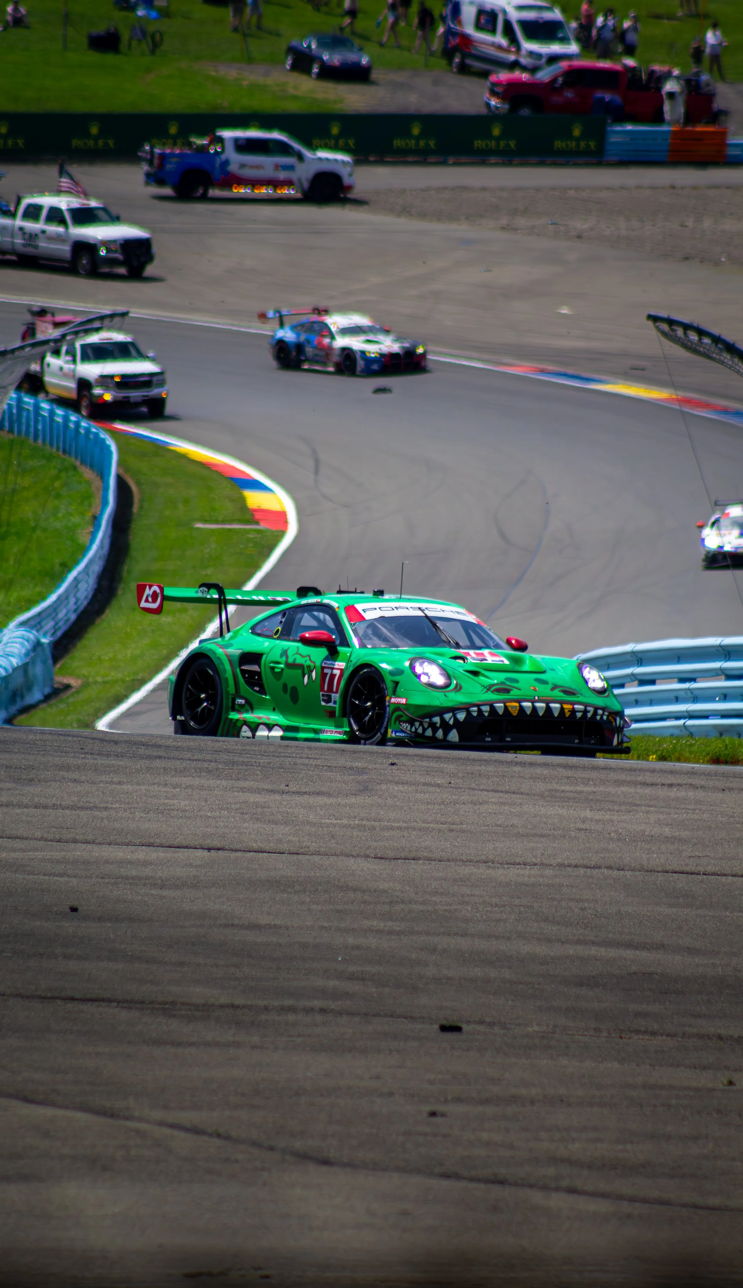 Green race car with cartoonish monster design on a racing track, with other vehicles and spectators in the background.