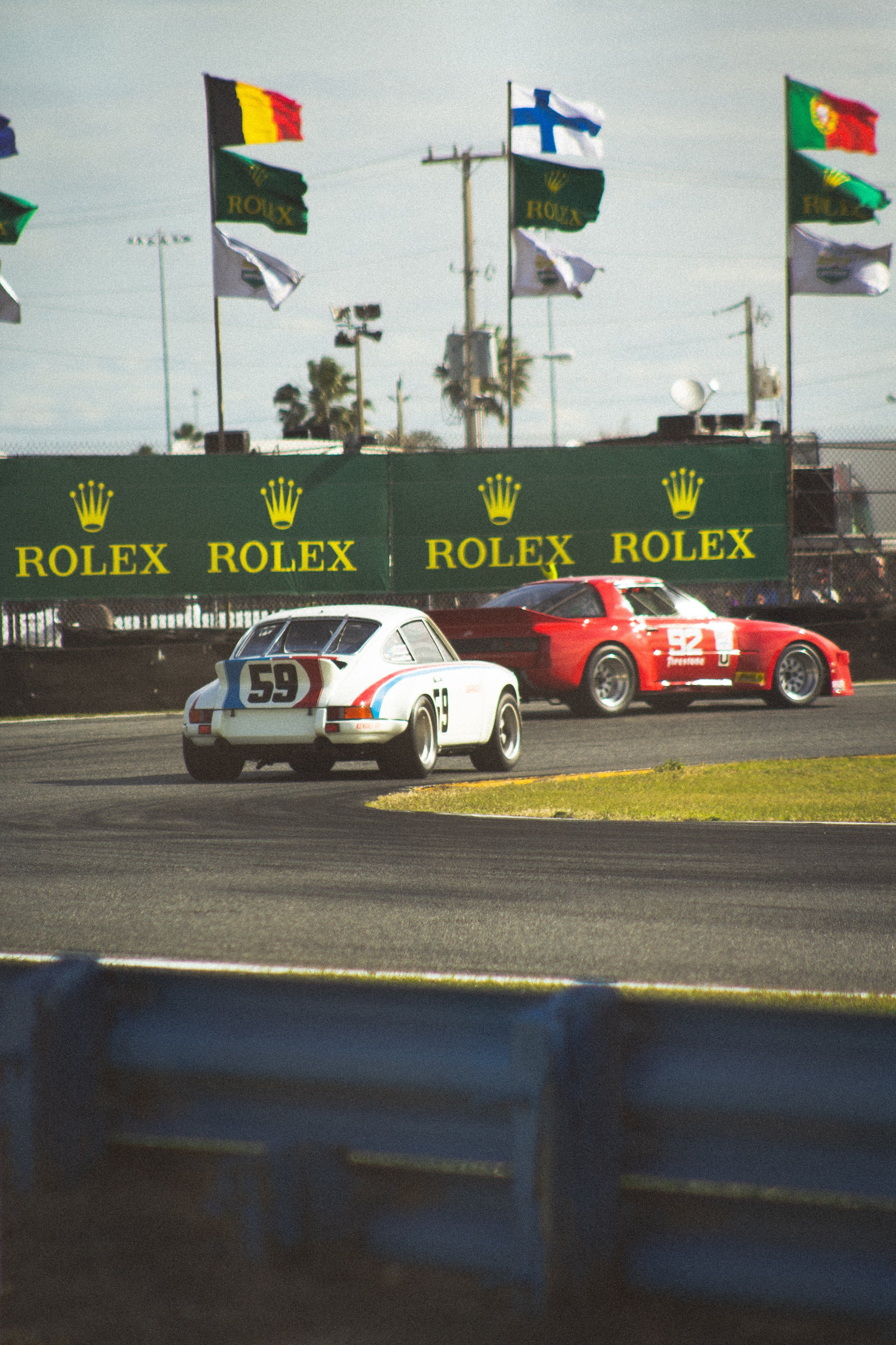 Two race cars on a track with flags and Rolex banners in the background.