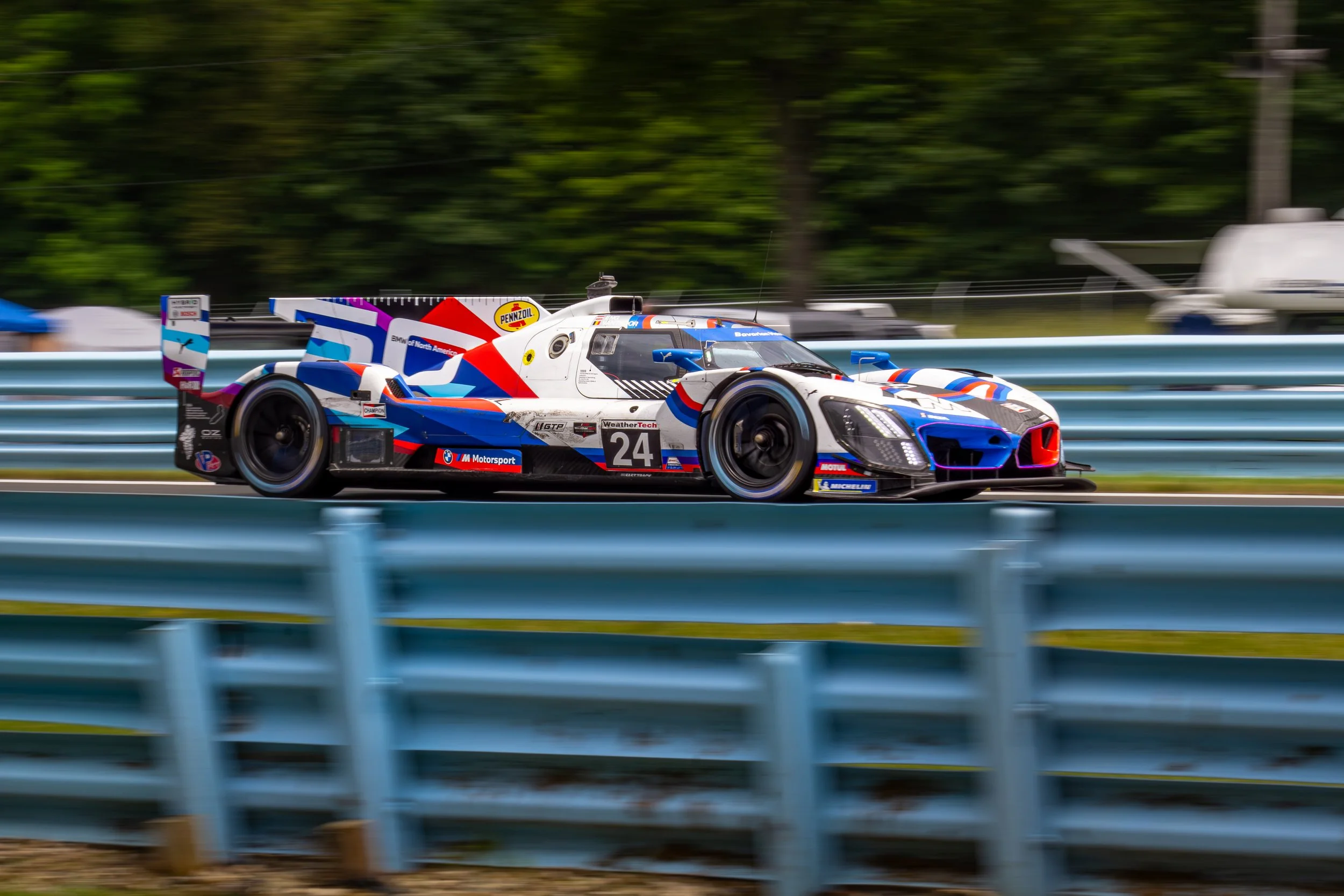 A racing car moving at high speed on a race track, with a background of trees and guardrails.