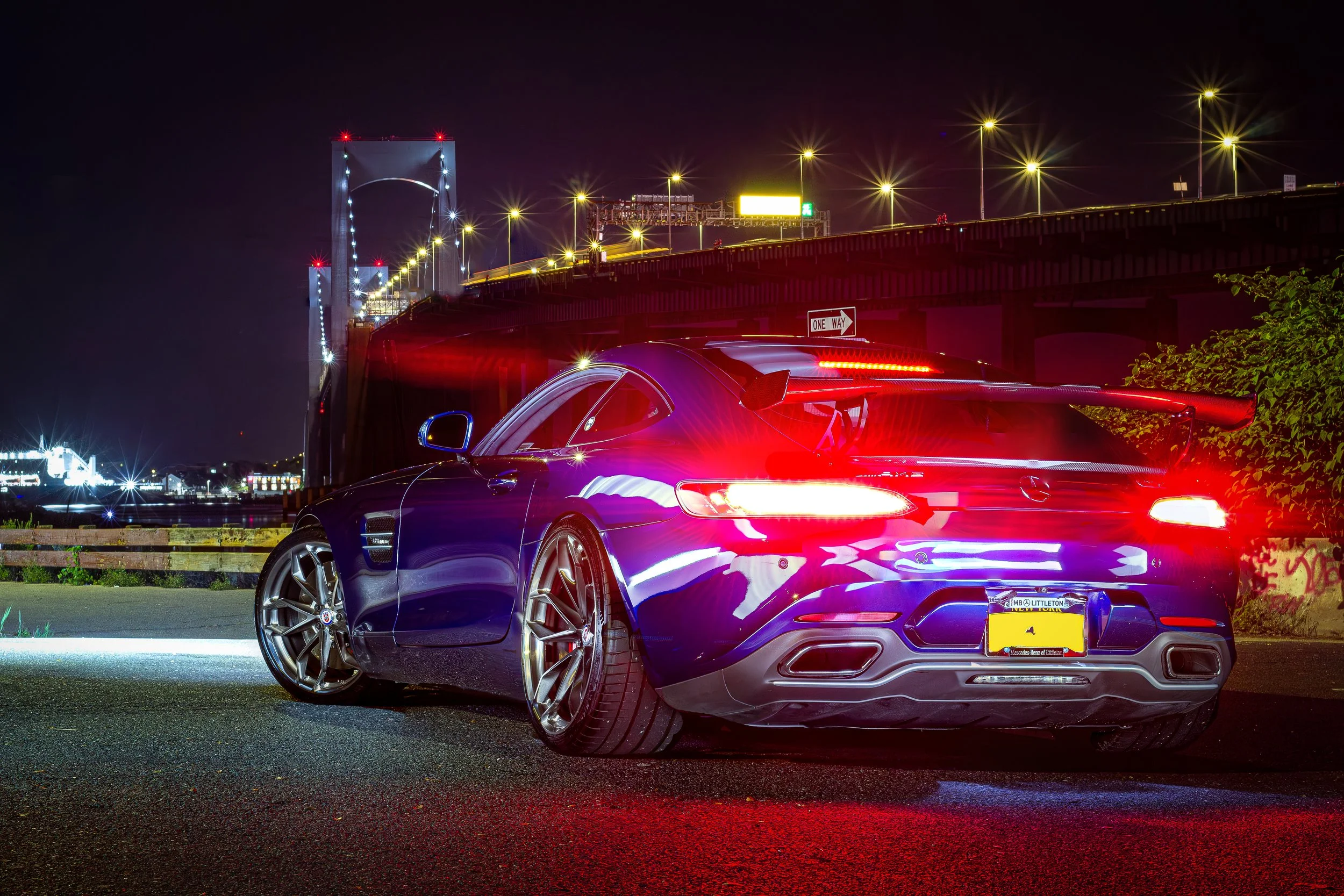 A dark blue sports car with bright taillights parked at night under a bridge, with city lights and a bridge illuminated in the background.