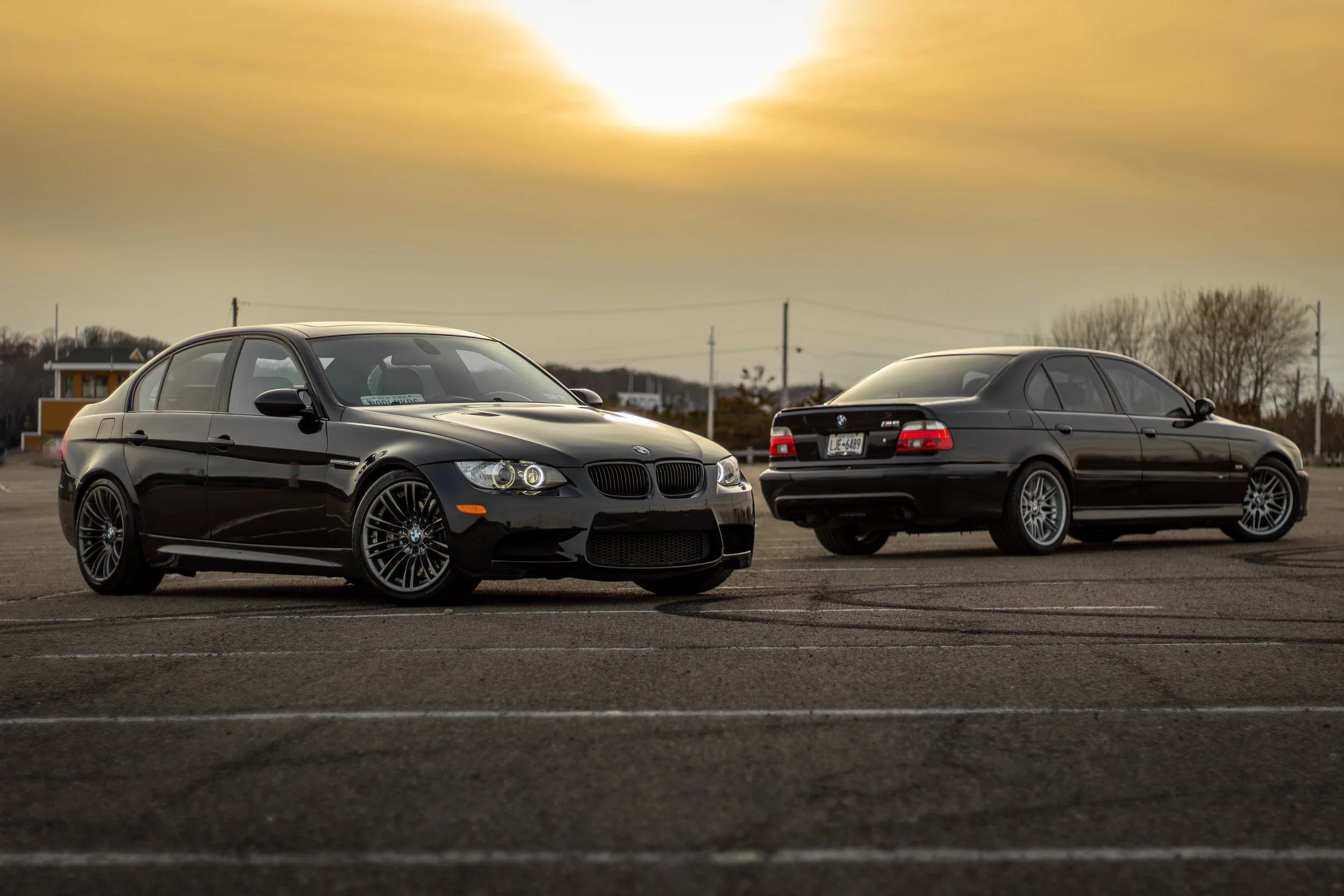 Two black BMW sedans parked in an empty parking lot during sunset with a yellow-orange sky.