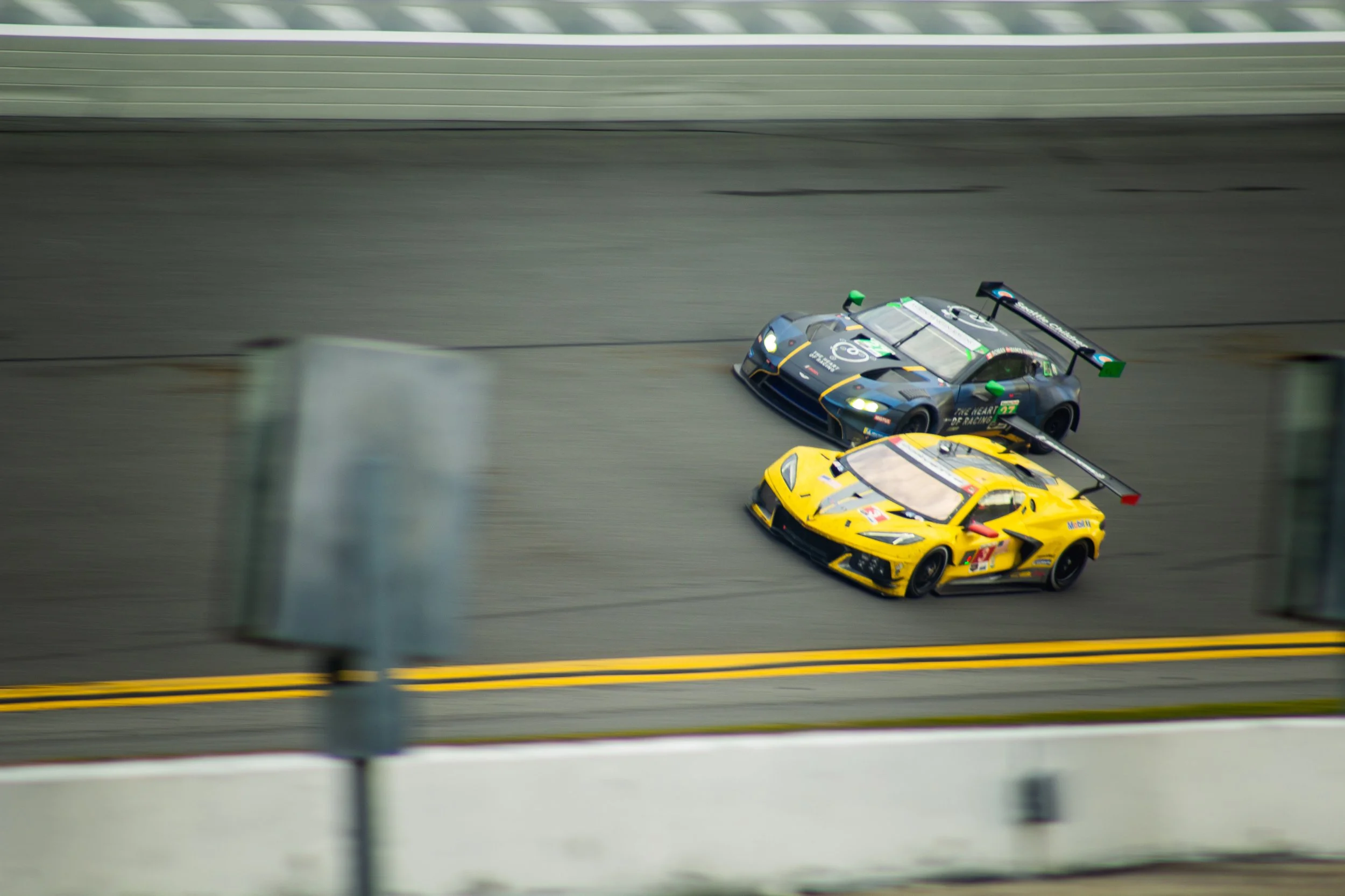 Two race cars on a track, one yellow and one dark gray, speeding around a corner with blurred barriers in the foreground.