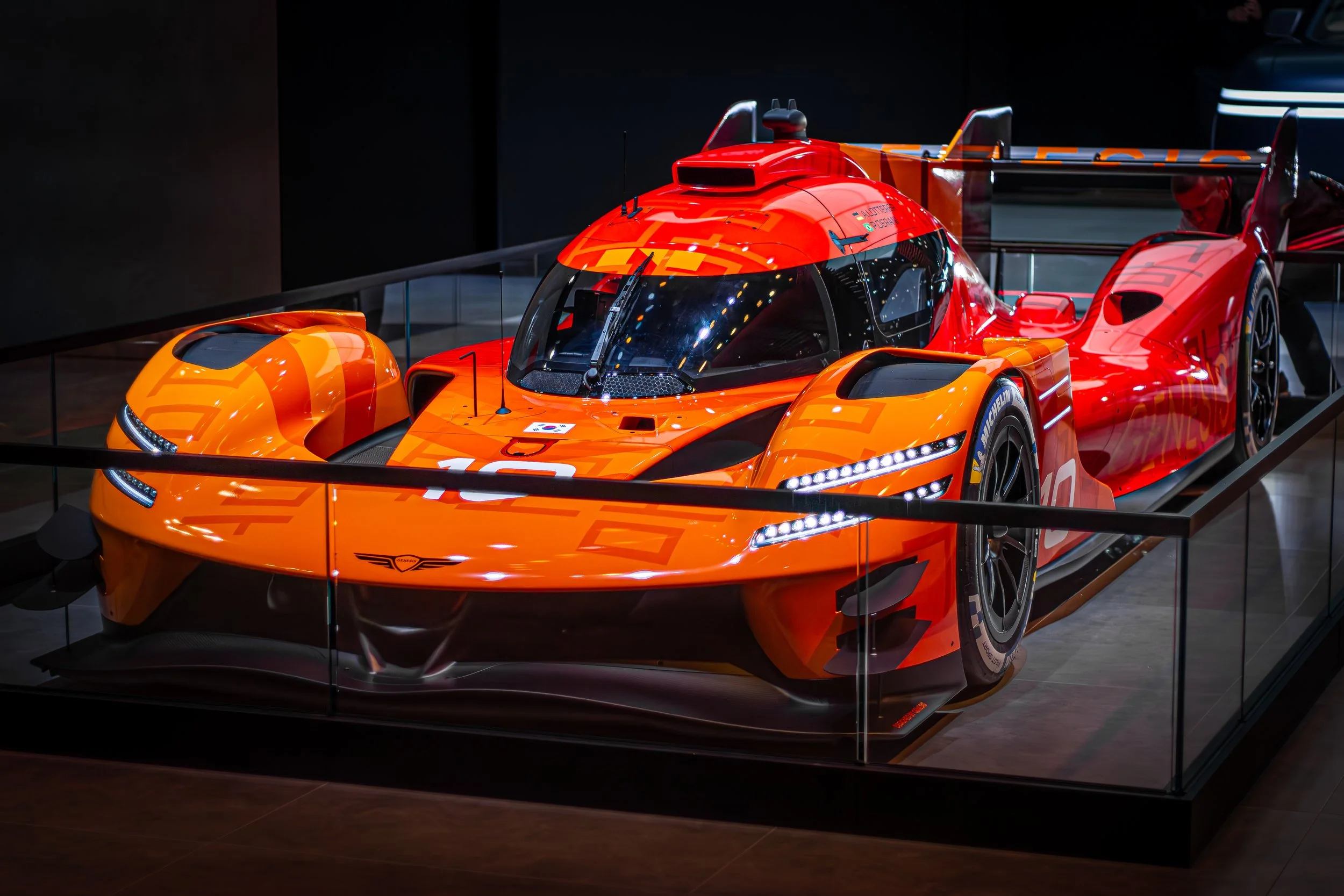 A bright orange Audi racing car displayed in a glass case at a museum, featuring aerodynamic design, a large rear wing, and Michelin racing tires.