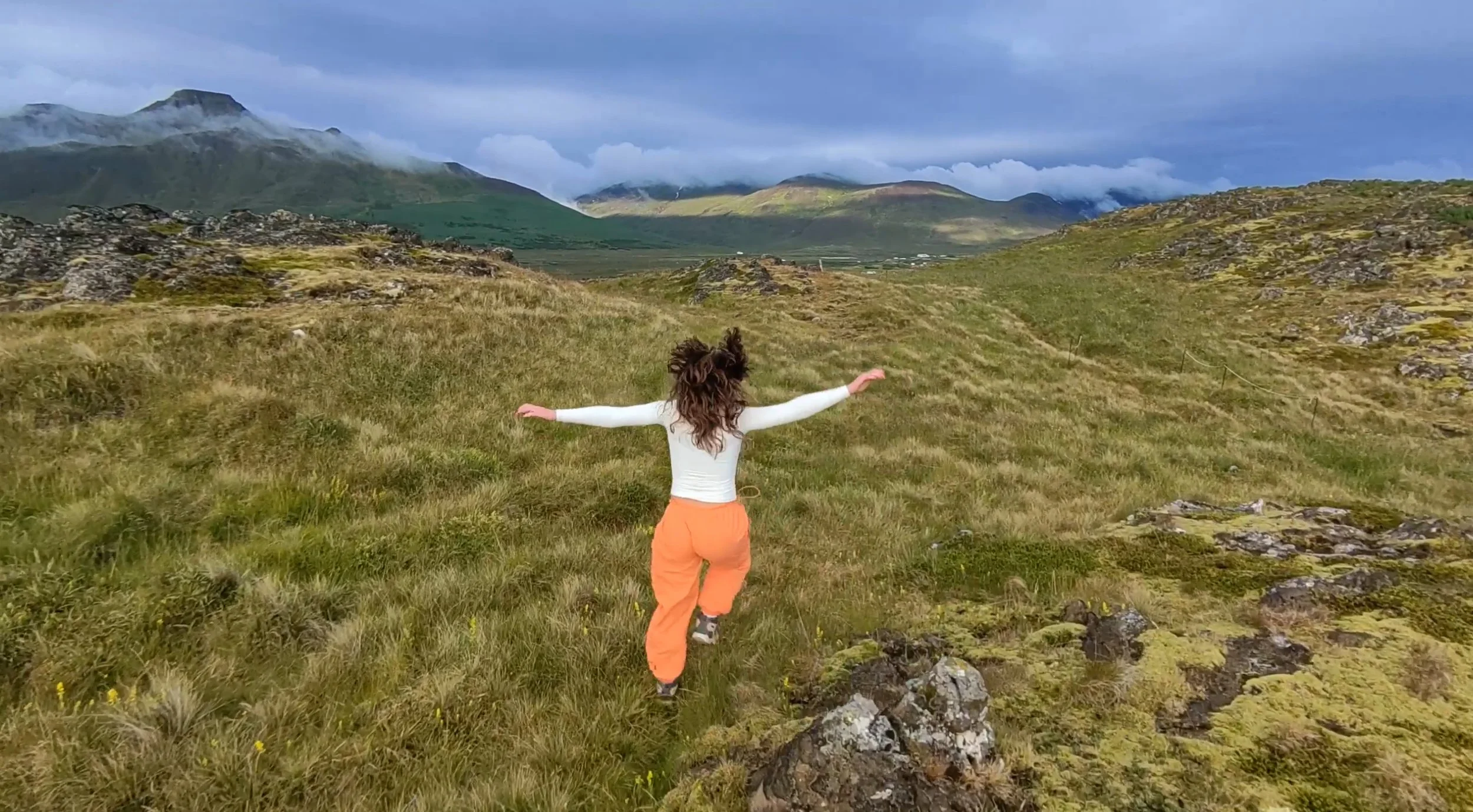 A person with curly hair and wearing a white top and orange pants is standing with arms outstretched on grassy terrain in a mountainous landscape with clouds covering the peaks.