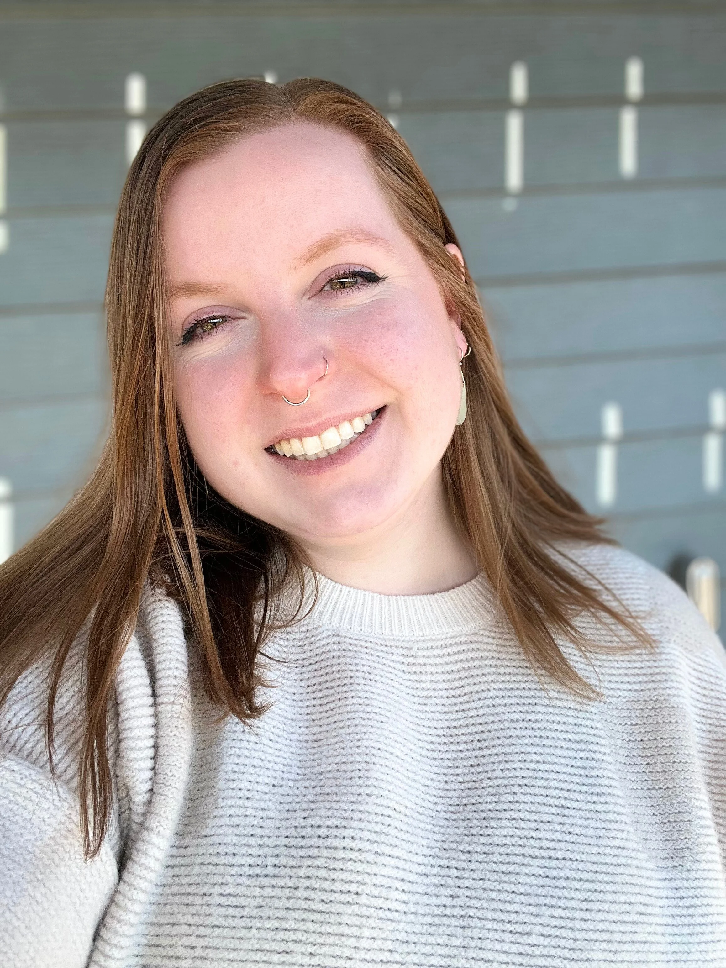 Photo of a red-headed, non-binary person, named Nat, smiling. They are wearing a creme colored sweater and have mid-length hair.