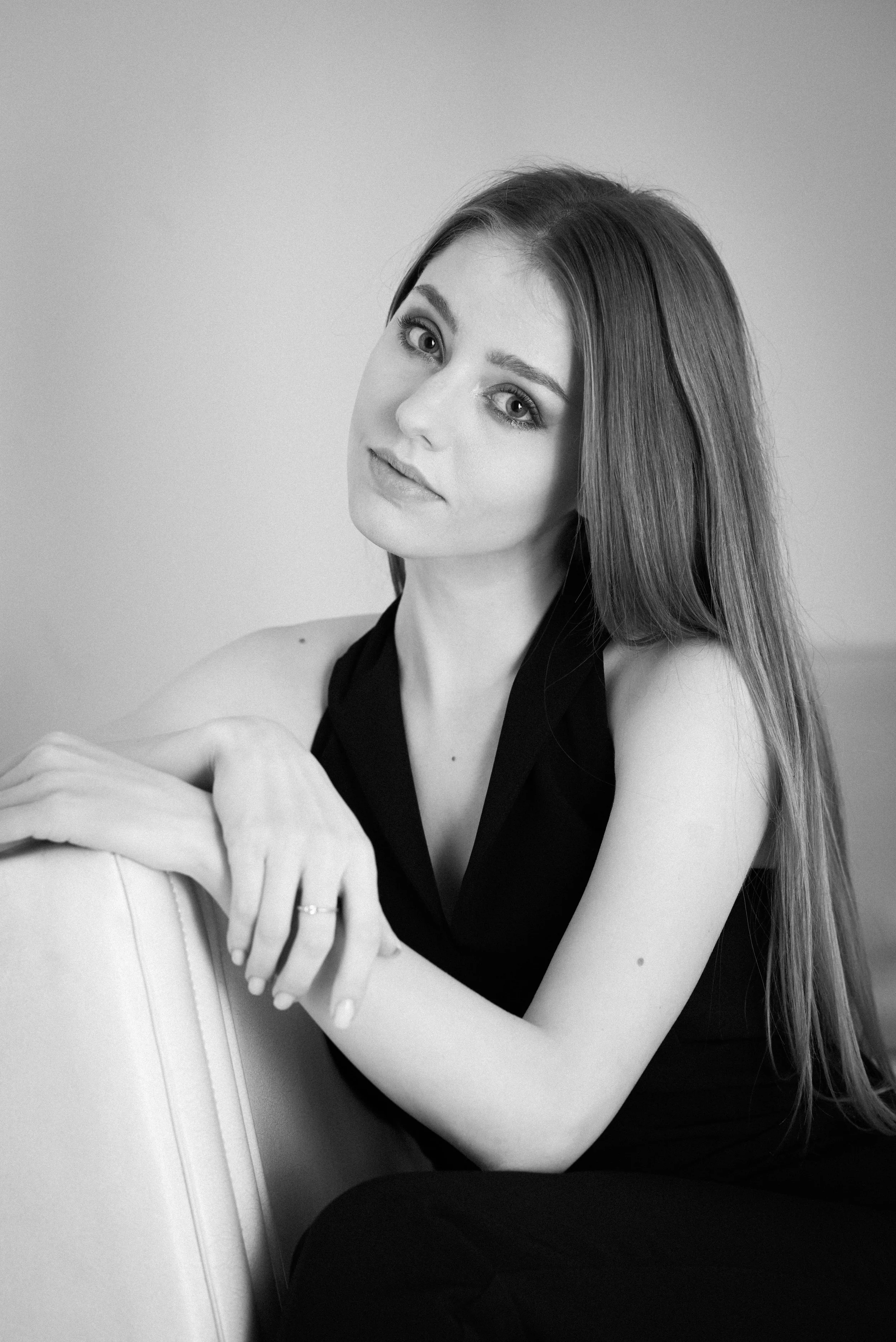 Black and white portrait of a young woman with long straight hair, sitting with her arms resting on a cushion, wearing a sleeveless black top, looking directly at the camera with a calm expression.