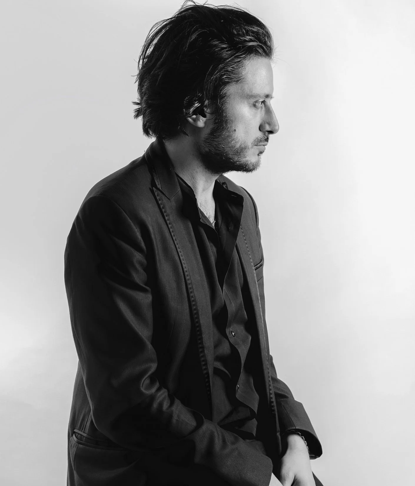 A black and white photo of a man with dark, wavy hair and a beard, wearing a dark suit jacket and shirt, sitting against a plain white background in profile view.