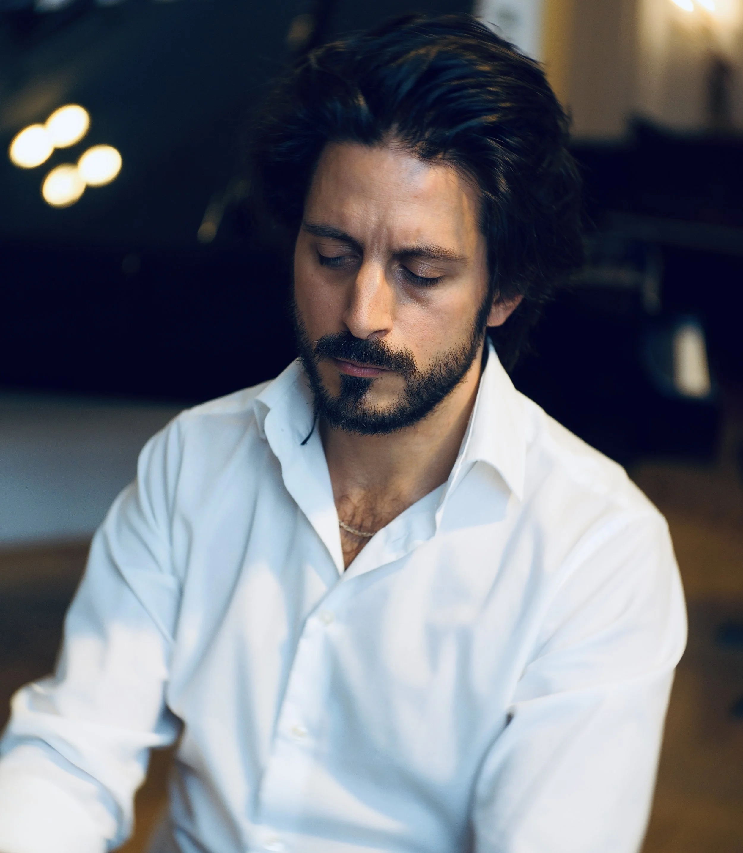 A man with dark hair, a beard, and a white shirt looks down thoughtfully in a dimly lit room.