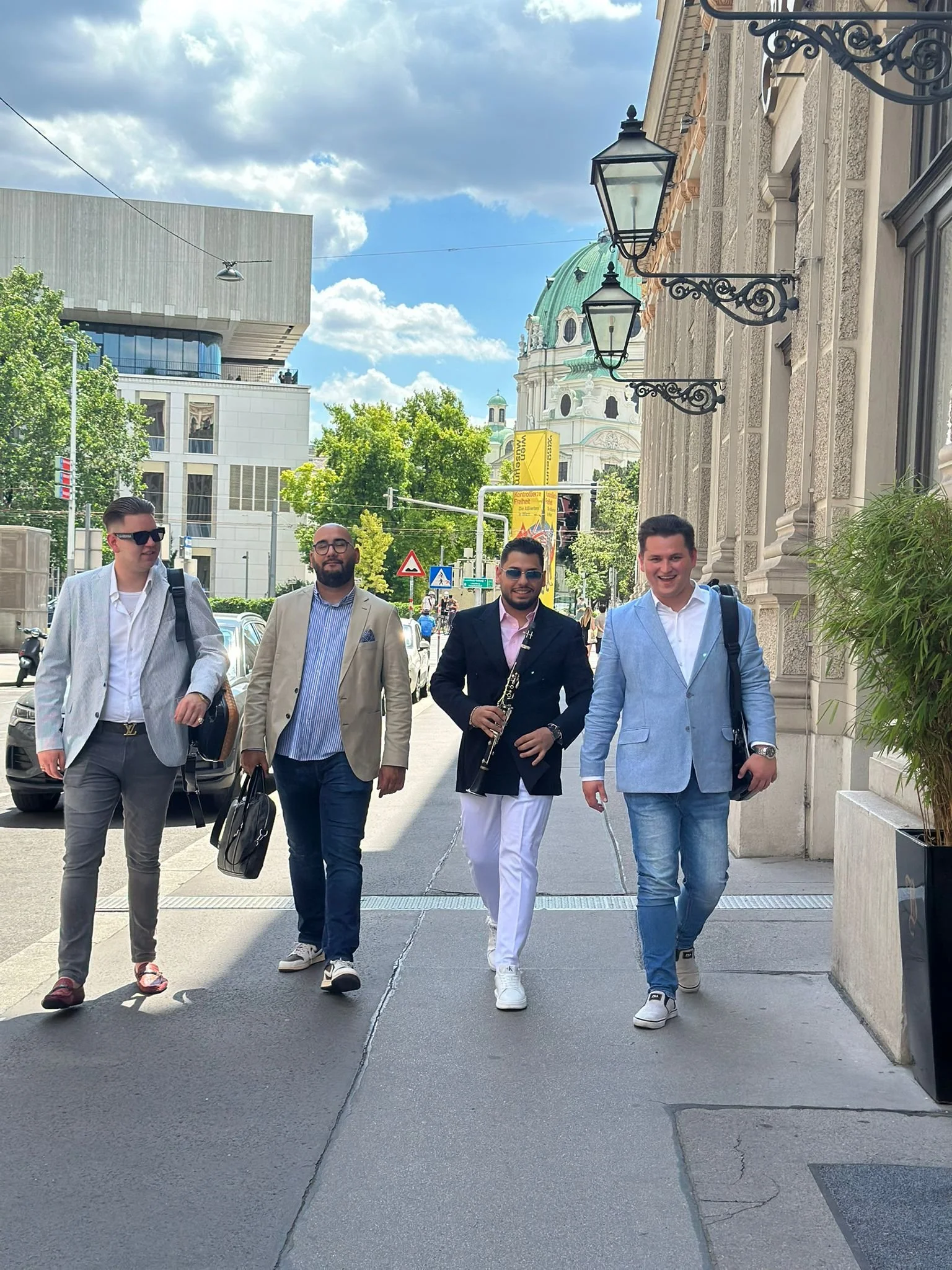 Four men walking down a city street, with one holding a clarinet, dressed in suits and casual outfits, with buildings and a church with a green dome in the background.