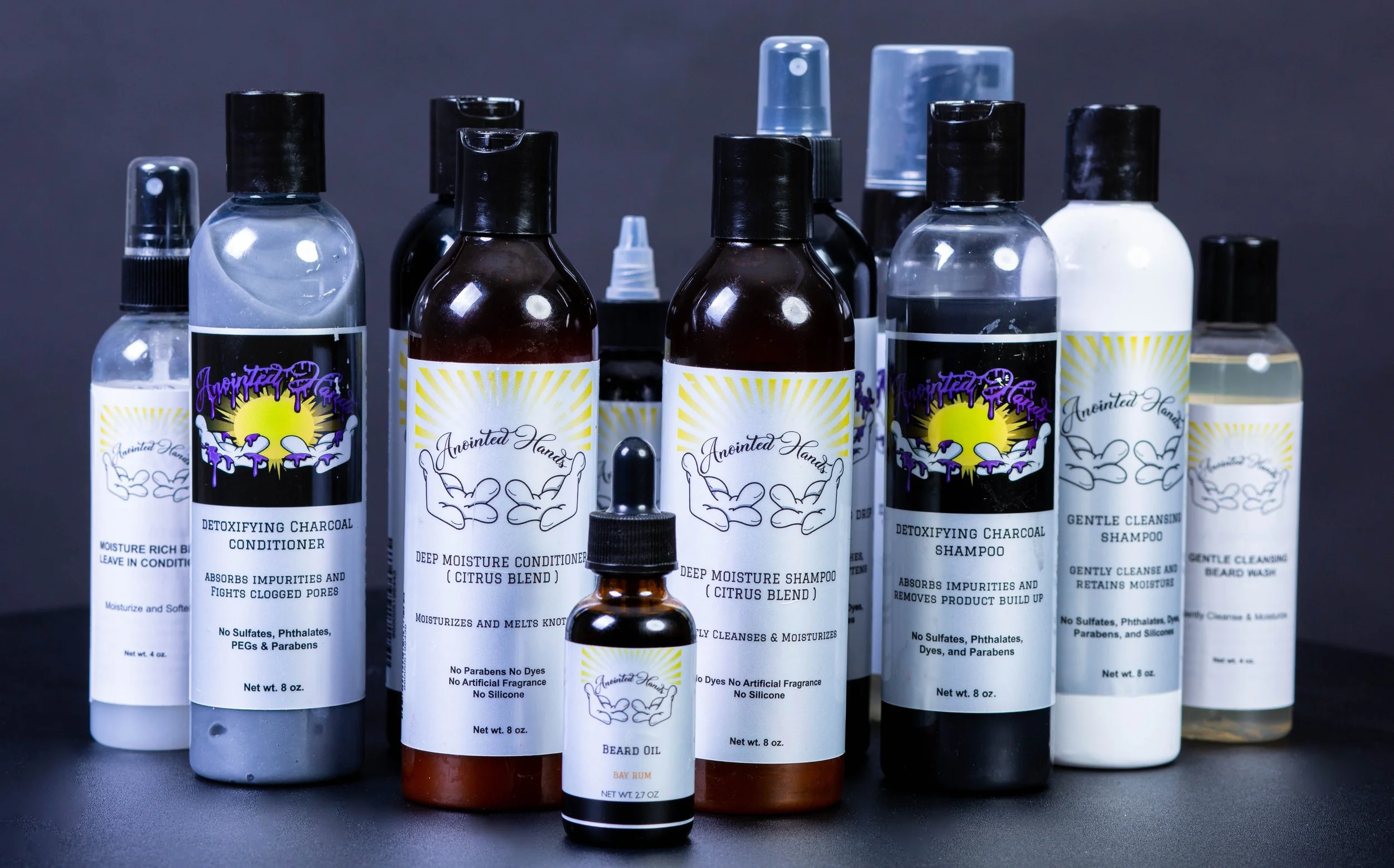 Collection of hair and beard care products including detoxifying charcoal conditioner, deep moisture conditioner, deep moisture shampoo, gentle cleansing shampoo, beard oil, and other bottles with labels indicating natural and sulfate-free ingredients.