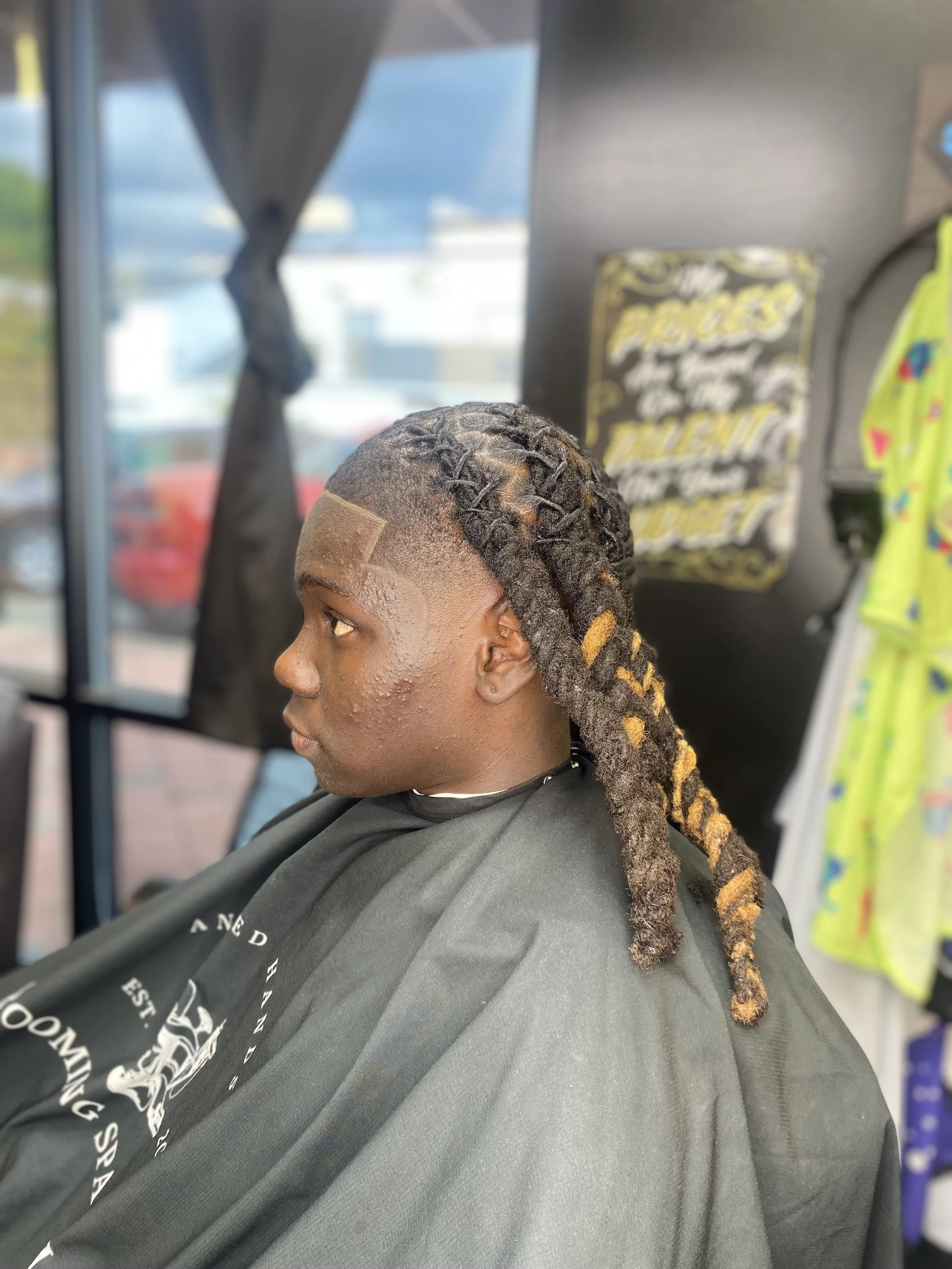 Young man with dreadlocks sitting in a barber shop chair, wearing a black barber cape, with a fresh haircut and styled dreadlocks. The shop has a window with curtains and a motivational sign in the background.