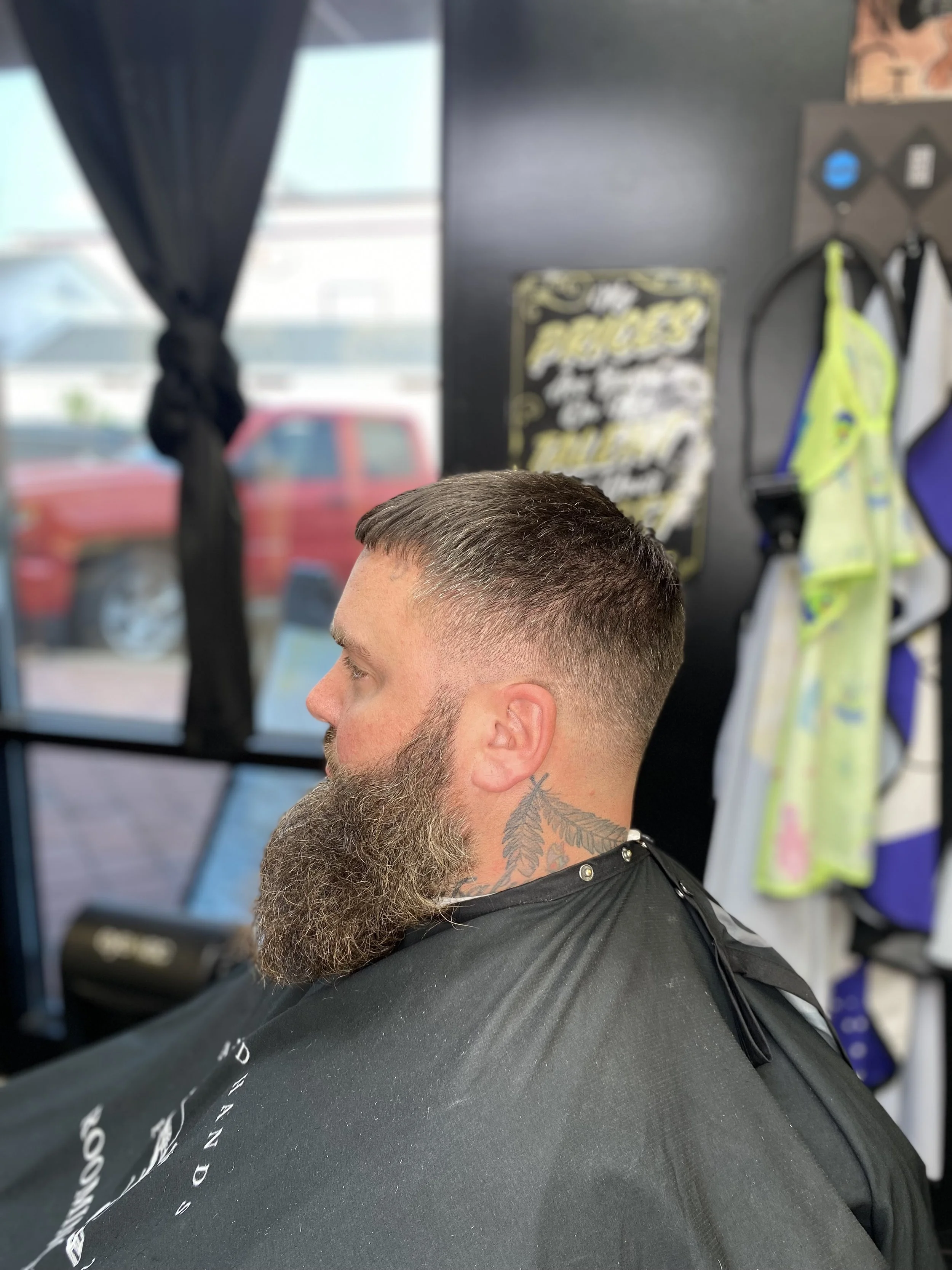 Side view of a man with short hair and a full beard sitting in a barber's chair, wearing a black cape, inside a barbershop with various tools and clothing hanging on the wall behind him.