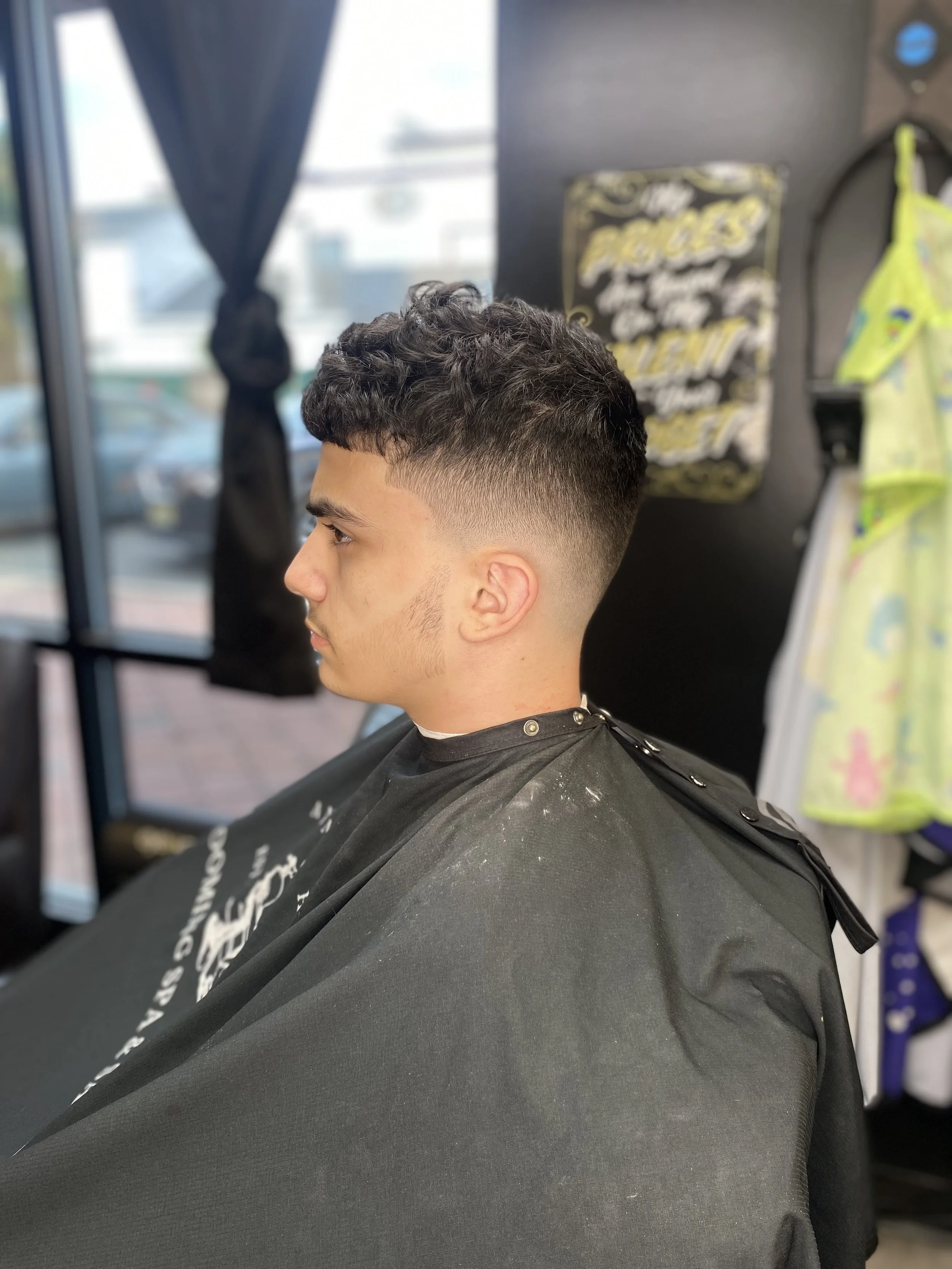 Young man with a textured haircut sitting in a barber shop, looking to the left, wearing a black cape. The barber shop has a window with black curtains and various items hanging on the wall.