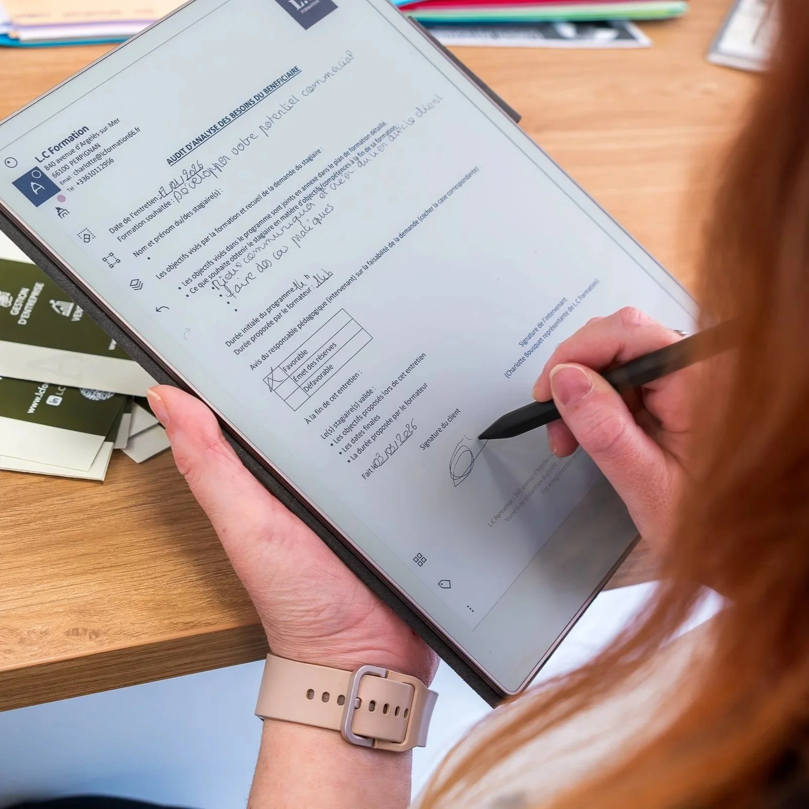 Une personne colorée en rouge ou orange écrit une signature manuscrite sur un document numérique présenté sur une tablette. La main de la personne tient un stylo ou un stylet et porte une montre beige. Le document semble être un formulaire ou un contrat, avec des cases à cocher, une signature, et plusieurs lignes de texte.