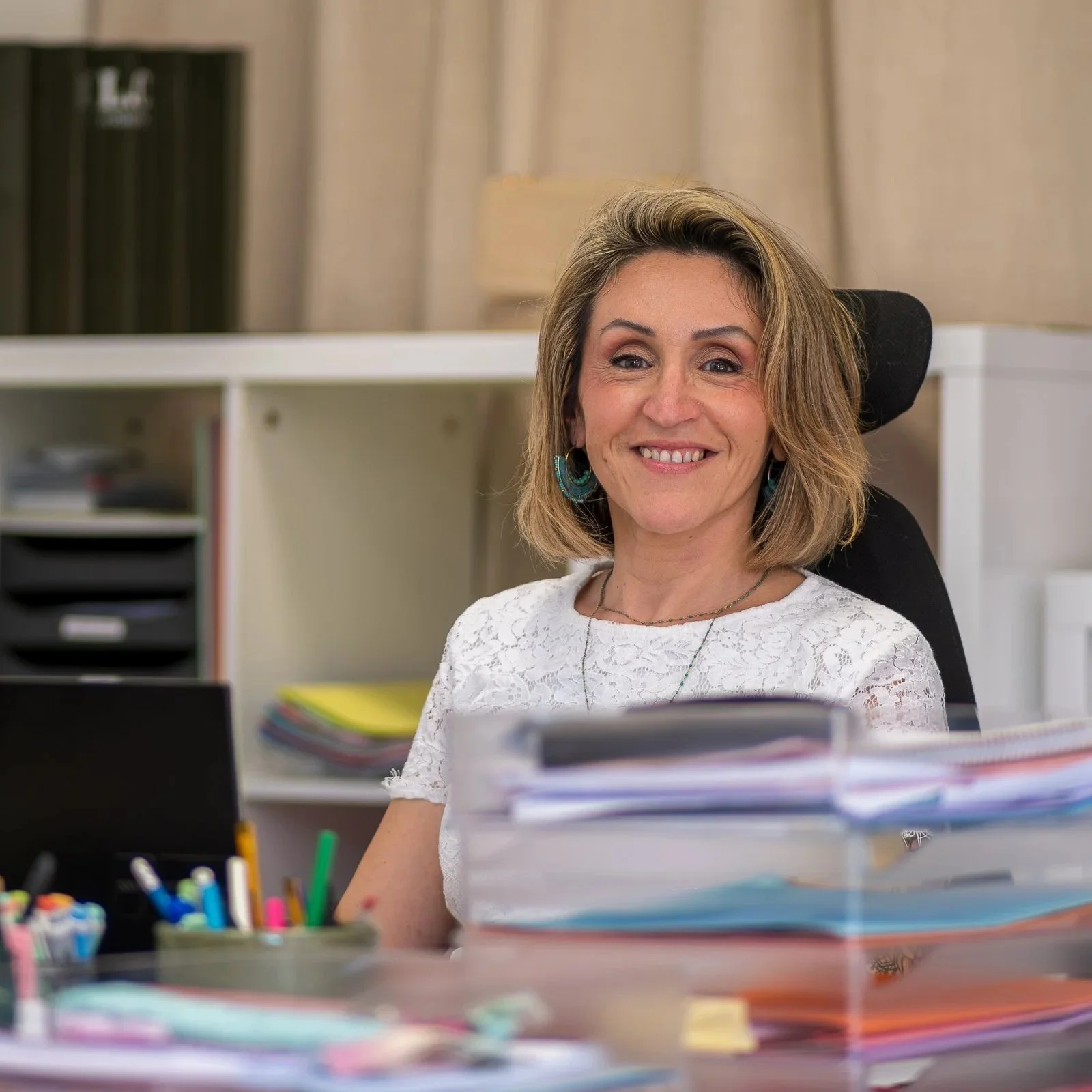 Mélanie Denizot, Assistante de direction L.C Formation. Une femme souriante assise à un bureau avec des étagères en arrière-plan et des piles de documents et fournitures de bureau sur le bureau.