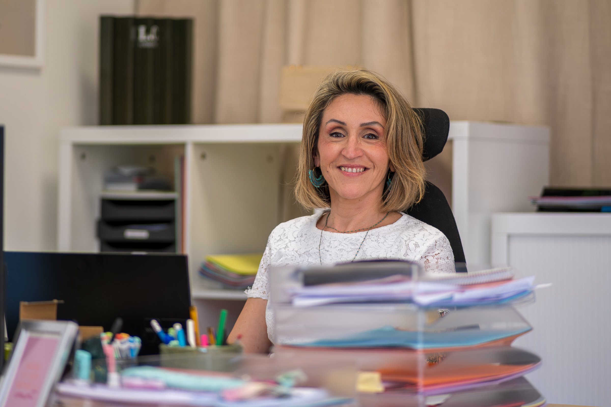 Mélanie Denizot, Assistante de direction L.C Formation. Une femme souriante assise à un bureau avec des étagères en arrière-plan et des piles de documents et fournitures de bureau sur le bureau.