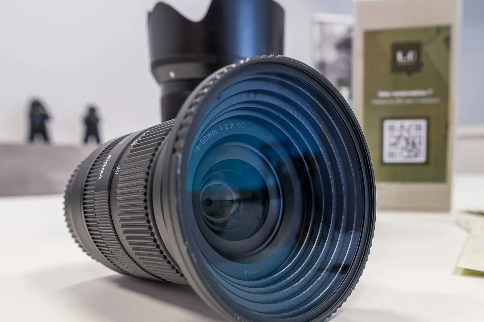 Objectif d'appareil photo de 18-50mm avec une lentille en verre, posé sur une surface blanche. En arrière-plan, un boîtier d'objectif et deux figurines sont visibles, ainsi qu'une boîte marron avec un QR code.