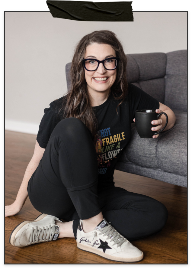 A woman with long wavy brown hair and glasses, sitting on a wooden floor, smiling at the camera, holding a black mug, wearing a black T-shirt and black pants, near a gray sofa in a room with white walls.