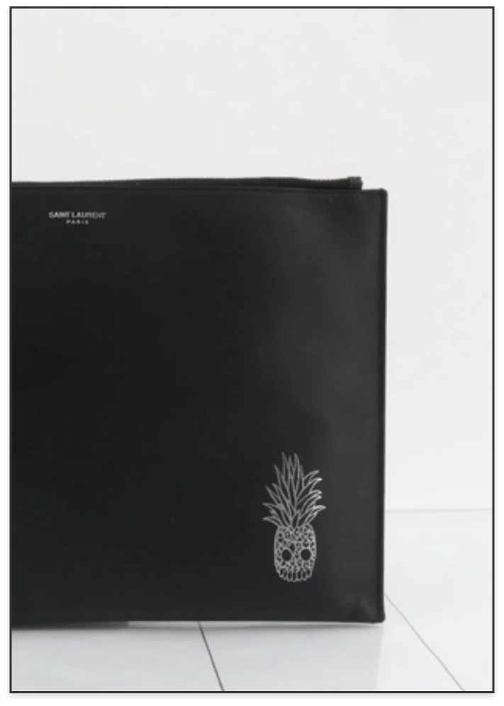 Black Yves Saint Laurent Paris clutch purse with a silver pineapple skull design in the bottom right corner, placed on a white tiled surface against a white background.