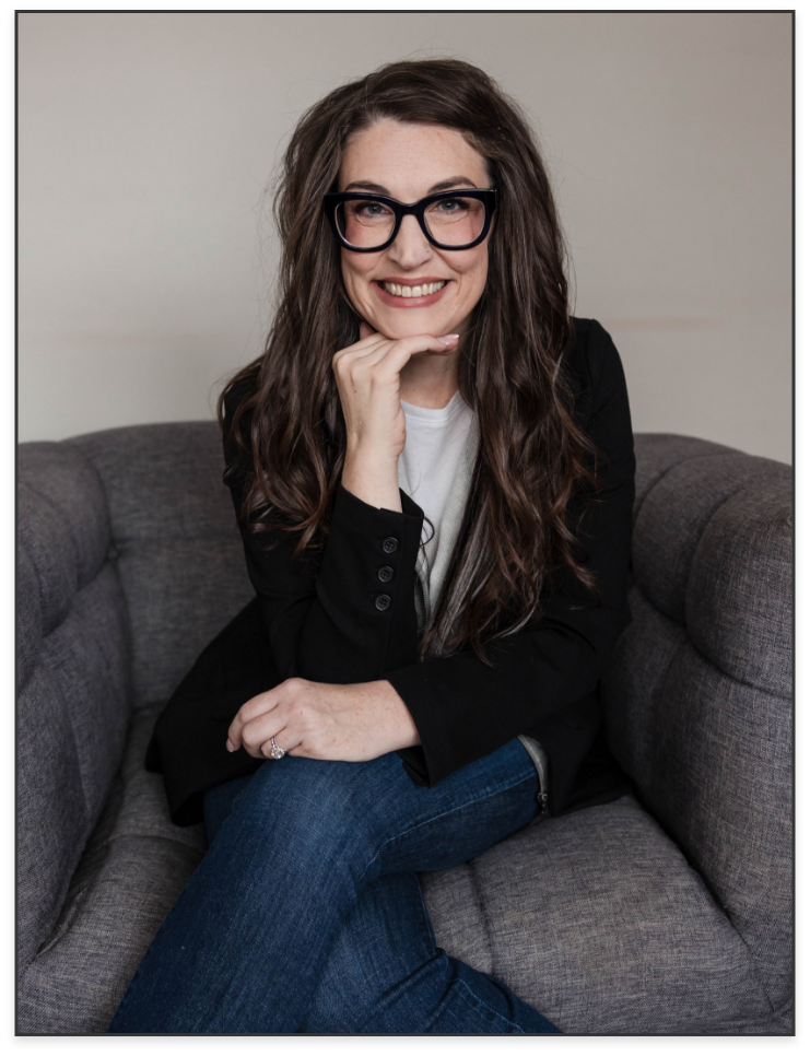 A woman with long, wavy brown hair wearing black glasses, a black blazer, and a white shirt, smiling and sitting on a gray couch.