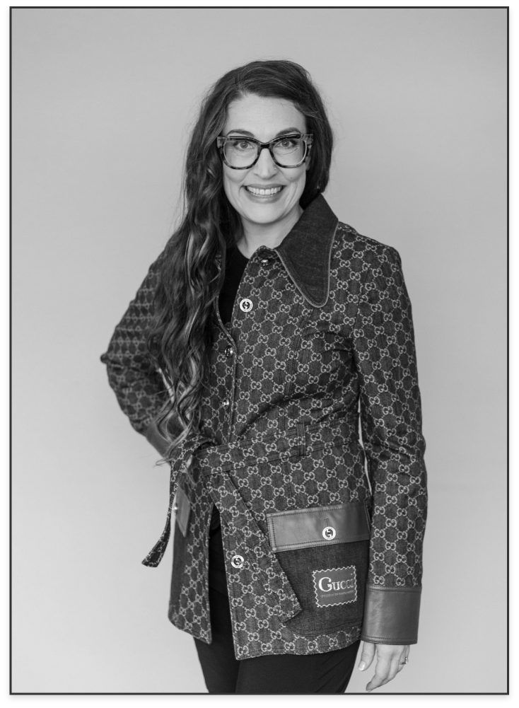 A woman with long wavy hair and glasses smiles at the camera against a plain background. She wears a Gucci patterned jacket with leather accents, pocket, and a belt, and black pants.