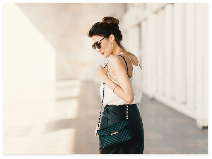 A woman with dark hair in a bun, wearing sunglasses, a white sleeveless top, and a black skirt, carrying a small green quilted purse on a silver chain, standing outdoors during daytime.