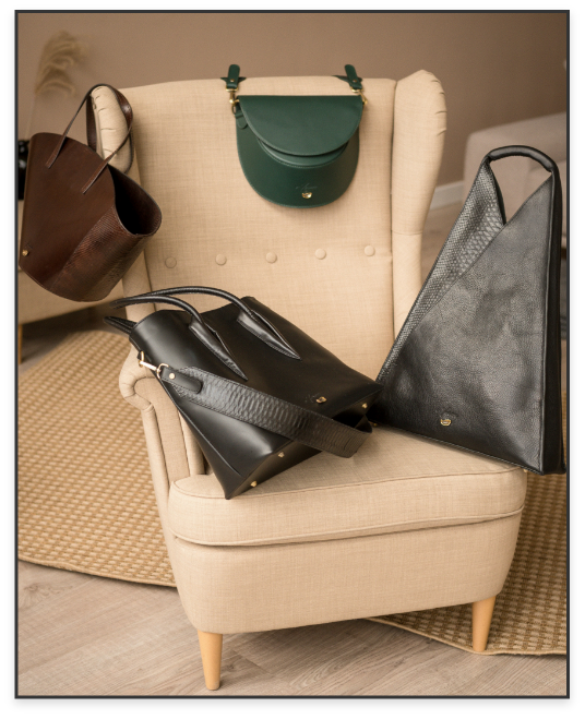A beige armchair with five handbags hanging and resting on and around it, including a brown tote, a green clutch, a black shoulder bag, a black large tote, and a textured gray bag.