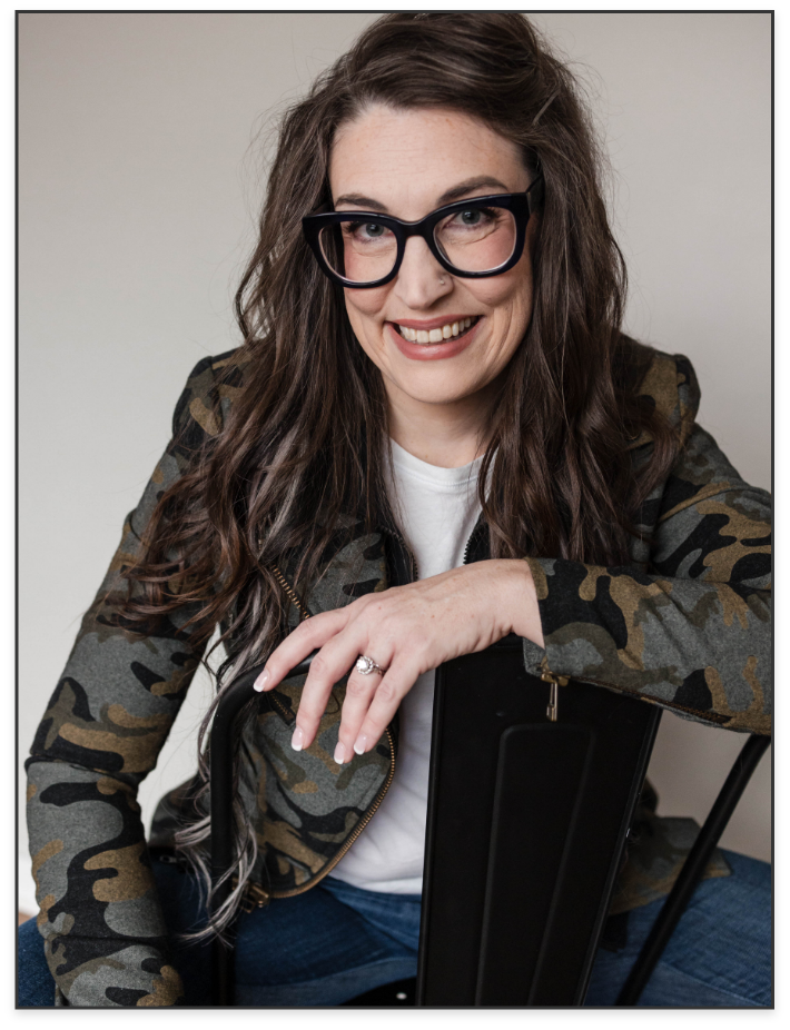 A woman with long, wavy brown hair, wearing black glasses, a camouflage jacket, and a white t-shirt, smiling and sitting on a black chair against a plain background.