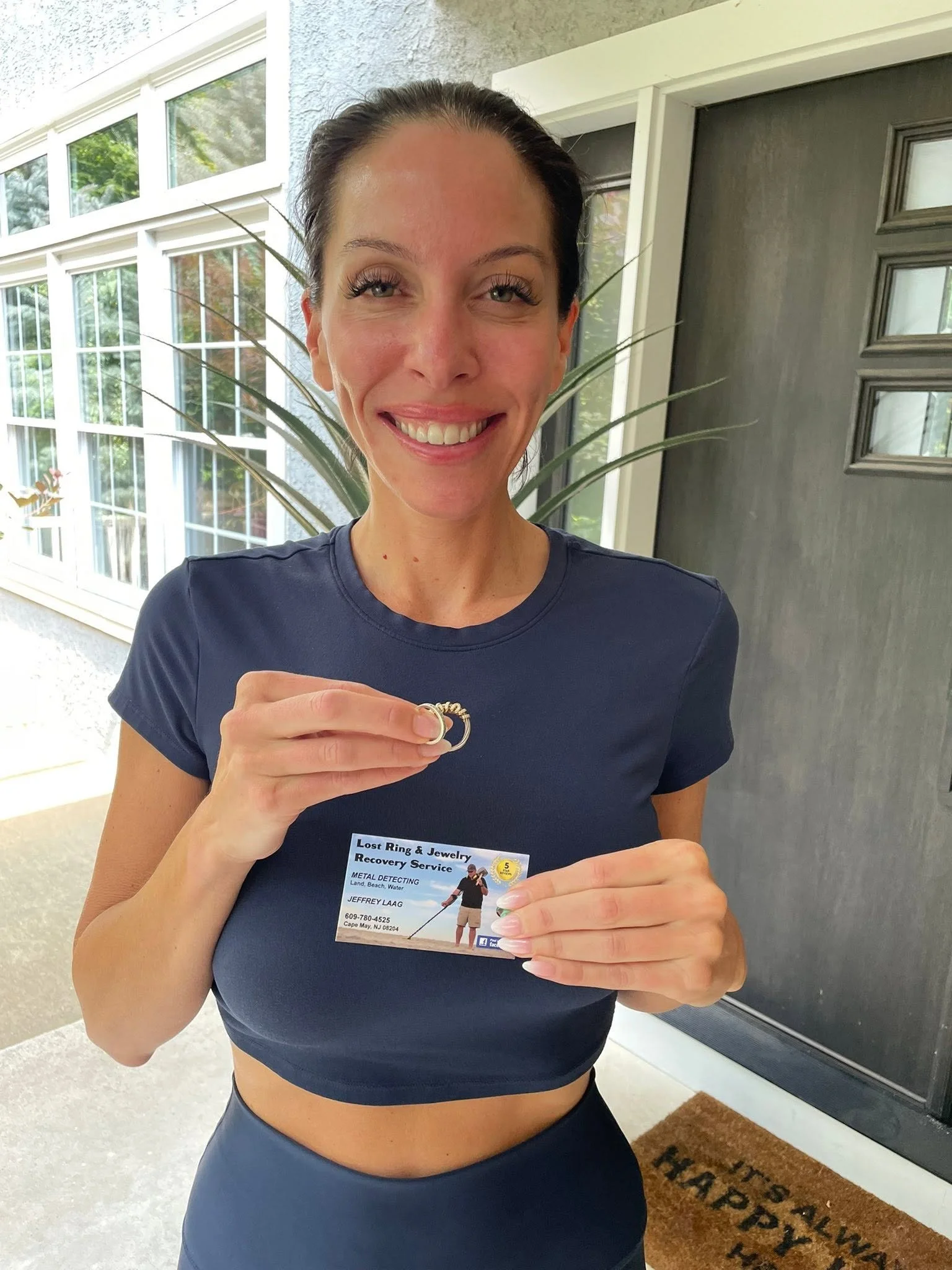 A woman is standing outside in front of a house, smiling and holding her recovered wedding rings and a business card that reads 'Lost Ring & Jewelry Recovery Service' with contact information. Rings recovered by Jeffrey Laag Ring Finders Cape May.