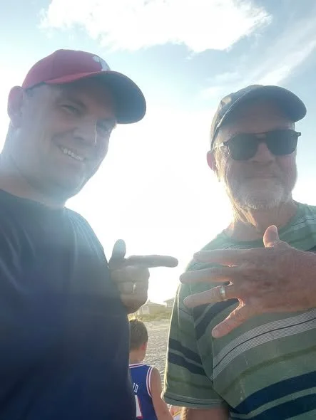 Two men smiling outdoors, one pointing at the wedding ring that was recovered by Ring Finders Cape May, with a child in the background and a blue sky with clouds.