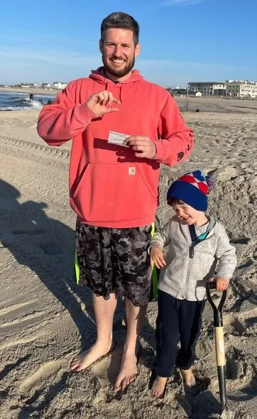 A man and a young boy on a sandy beach, with the man holding a piece of paper and the boy holding a small shovel. The man is wearing a red hoodie and patterned shorts, and the boy is wearing a jacket, hat, and dark pants. In the background, there are buildings and a clear blue sky.