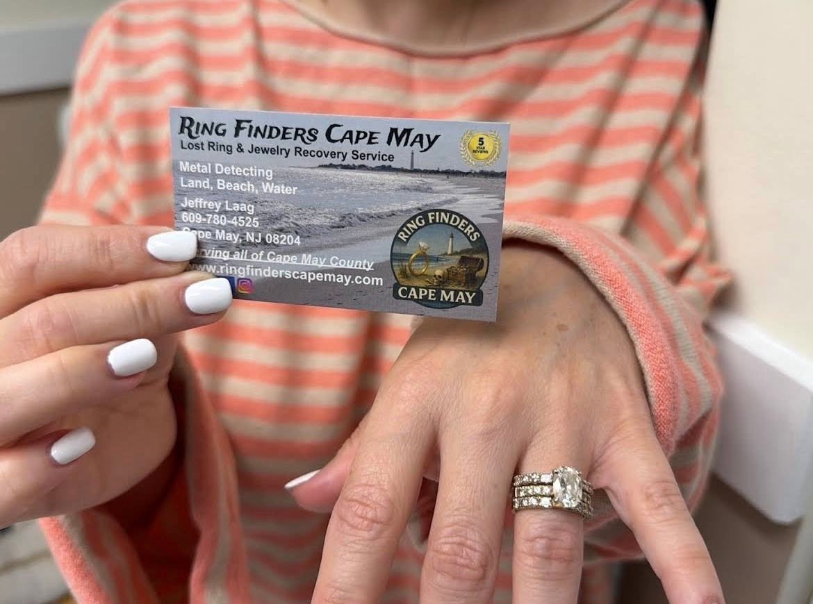 Person showing a business card for Ring Finders Cape May, holding up a ring on their finger with a large engagement ring and matching band. The person has a coral and white striped shirt and white nails.