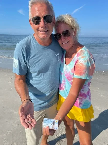 A man and woman stand together on a beach, both wearing sunglasses and smiling. The man is holding a recovered charm, and the woman is holding a ticket or card. The ocean and blue sky are visible in the background in Cape May New Jersey.