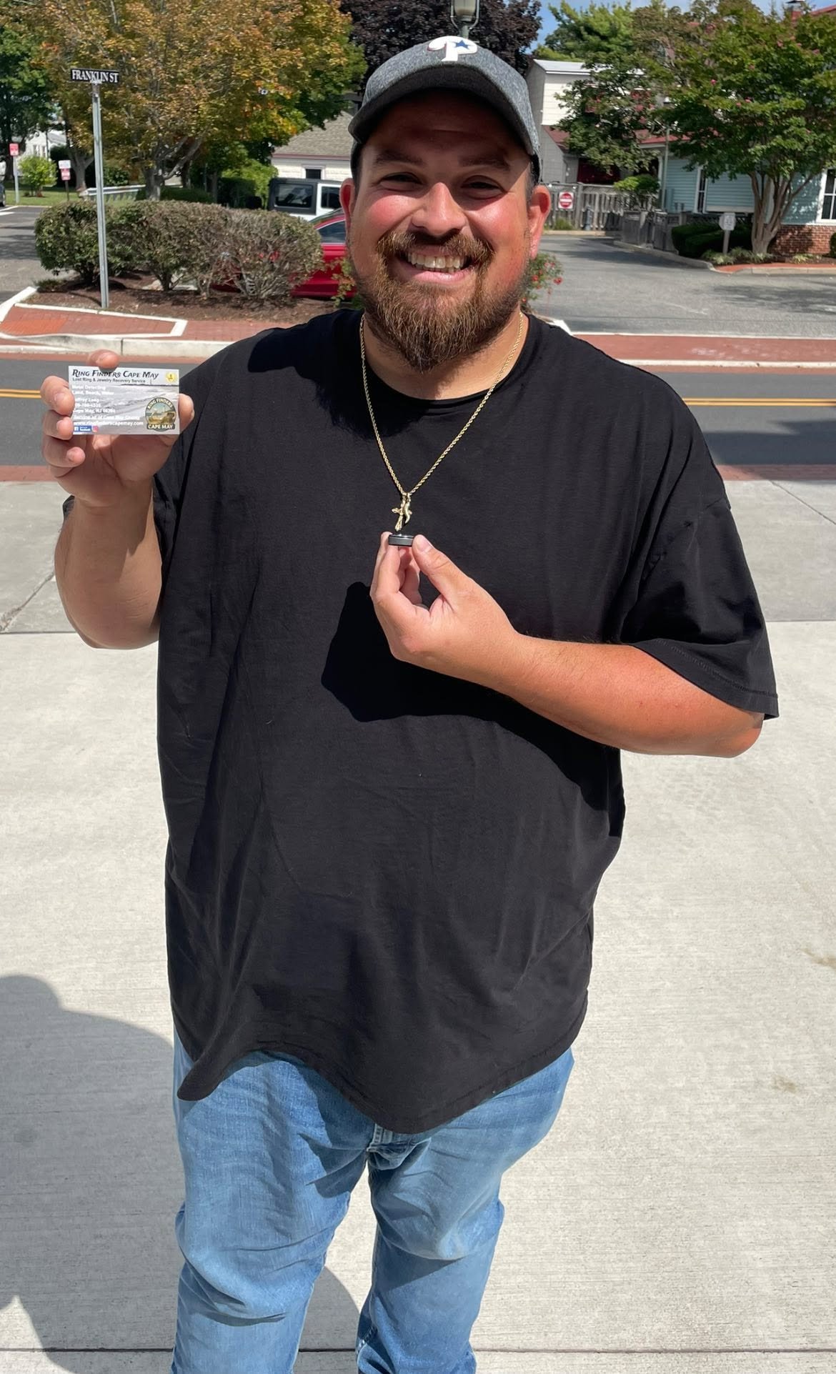 A smiling man with a beard and wearing a baseball cap holding a business card in one hand and a necklace in the other, standing on a sidewalk with trees and houses in the background.