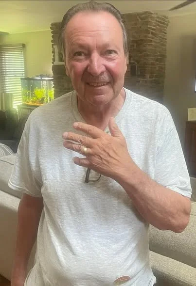 Older man with gray hair and beard, wearing a light gray T-shirt, smiling and touching his neck with his left hand, in a room with brick wall and aquarium in background.