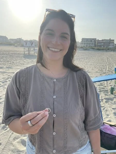 A woman smiling at the beach, holding a small object in her hand, with beach houses and sun in the background.