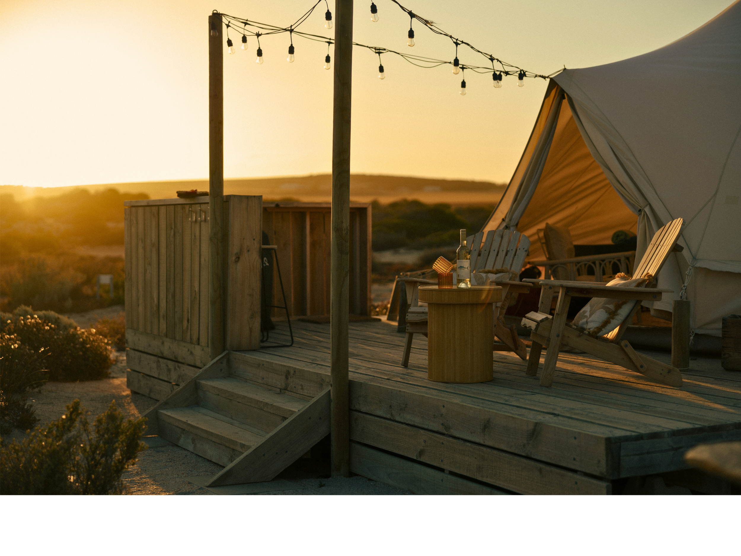 A wooden deck with Adirondack chairs, a small round table with a bottle and a drink, and a tent in the background during sunset. String lights hang above the deck, creating a cozy outdoor setting.