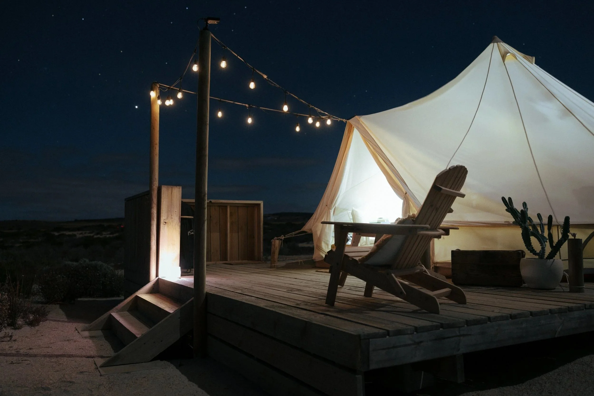 A nighttime scene of a campsite featuring a white canvas tent with wooden poles and a bench inside, illuminated by warm lighting, with string lights crossing above, set against a dark starry sky.
