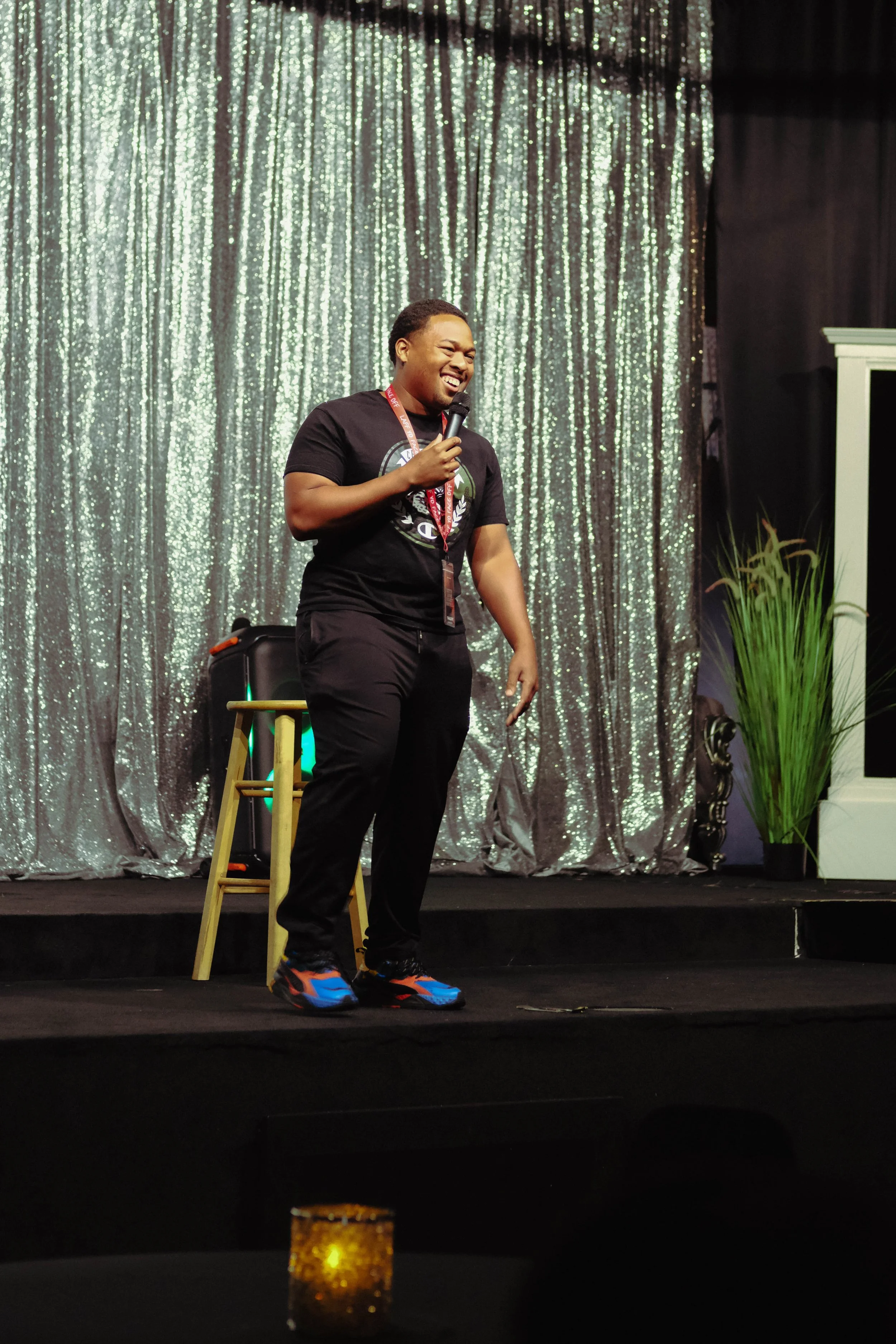 A person standing on a stage, smiling and holding a microphone. They are wearing black clothing and colorful sneakers. The background features a shiny, silver curtain, and there is a potted plant to the right. A small illuminated candle is visible in the foreground.