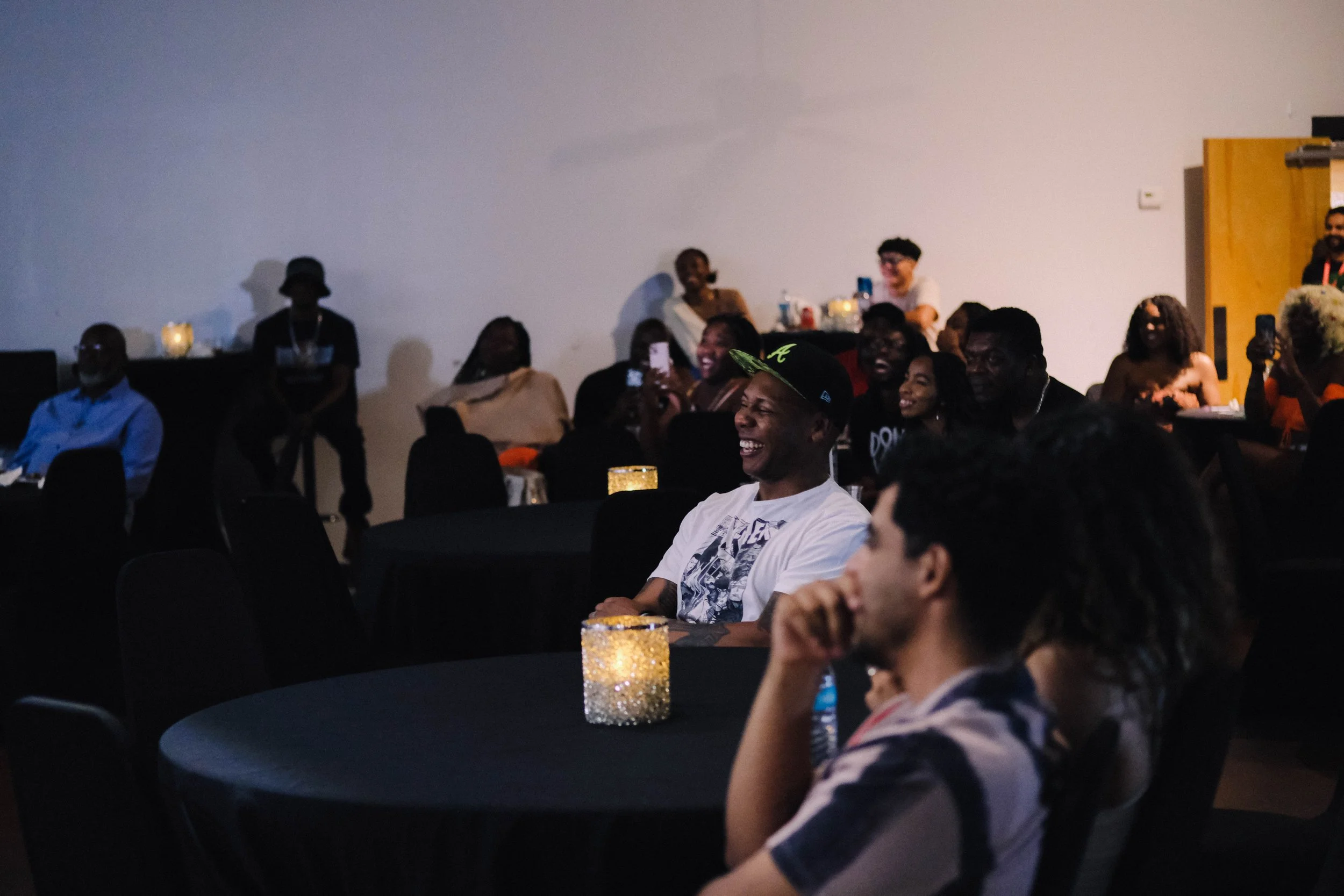 A group of people attending an event or gathering, sitting at tables with decorative candles, some smiling and taking photos, in a dimly lit room with plain walls.