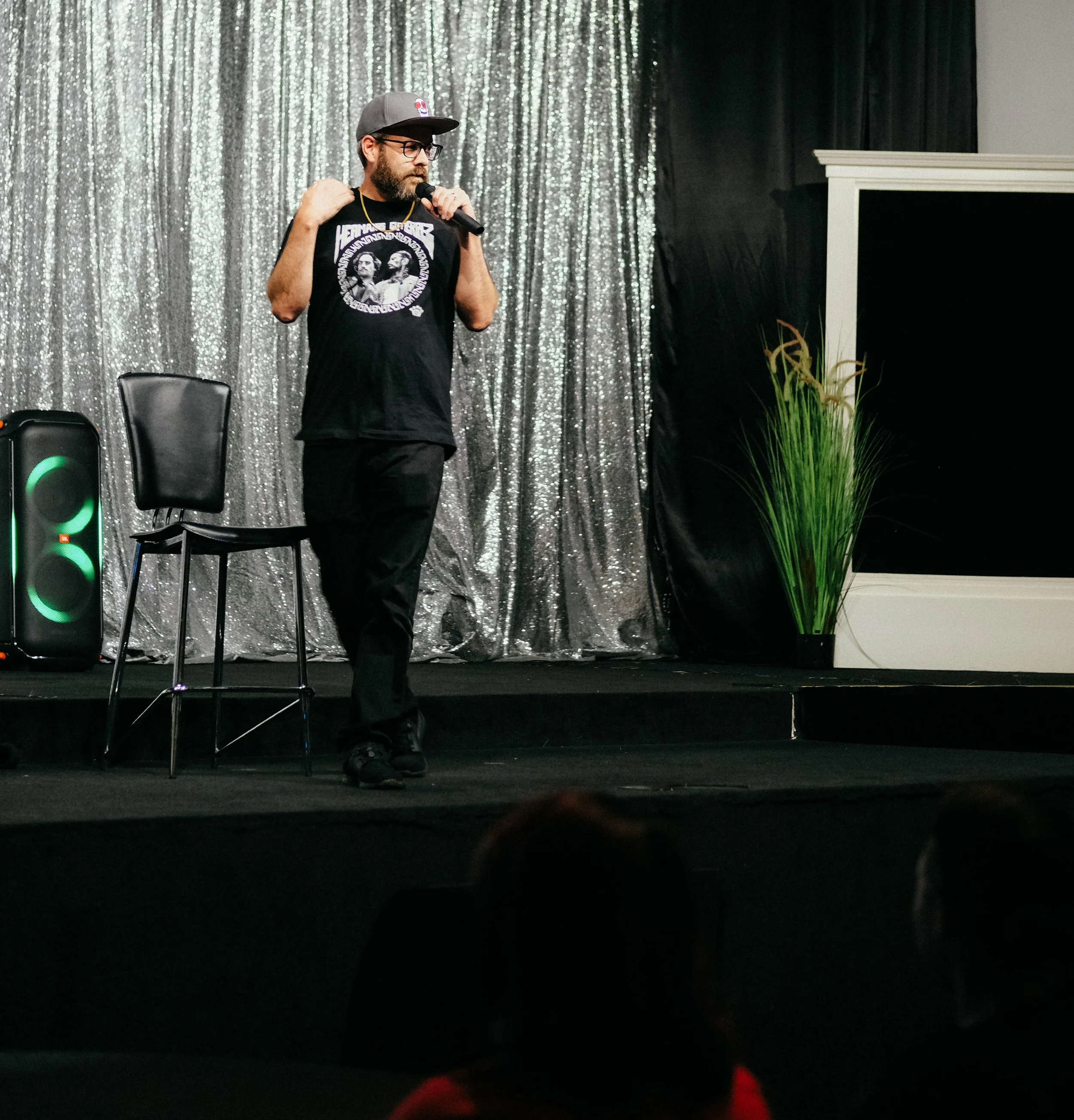 A man with glasses, a beard, and a dark t-shirt featuring monochrome images stands on a stage with a microphone. Behind him is a shiny, silver, sequin curtain, and to his right is a large black screen or panel. There is a black chair and a speaker next to him, and some plants are visible at the edge of the stage.