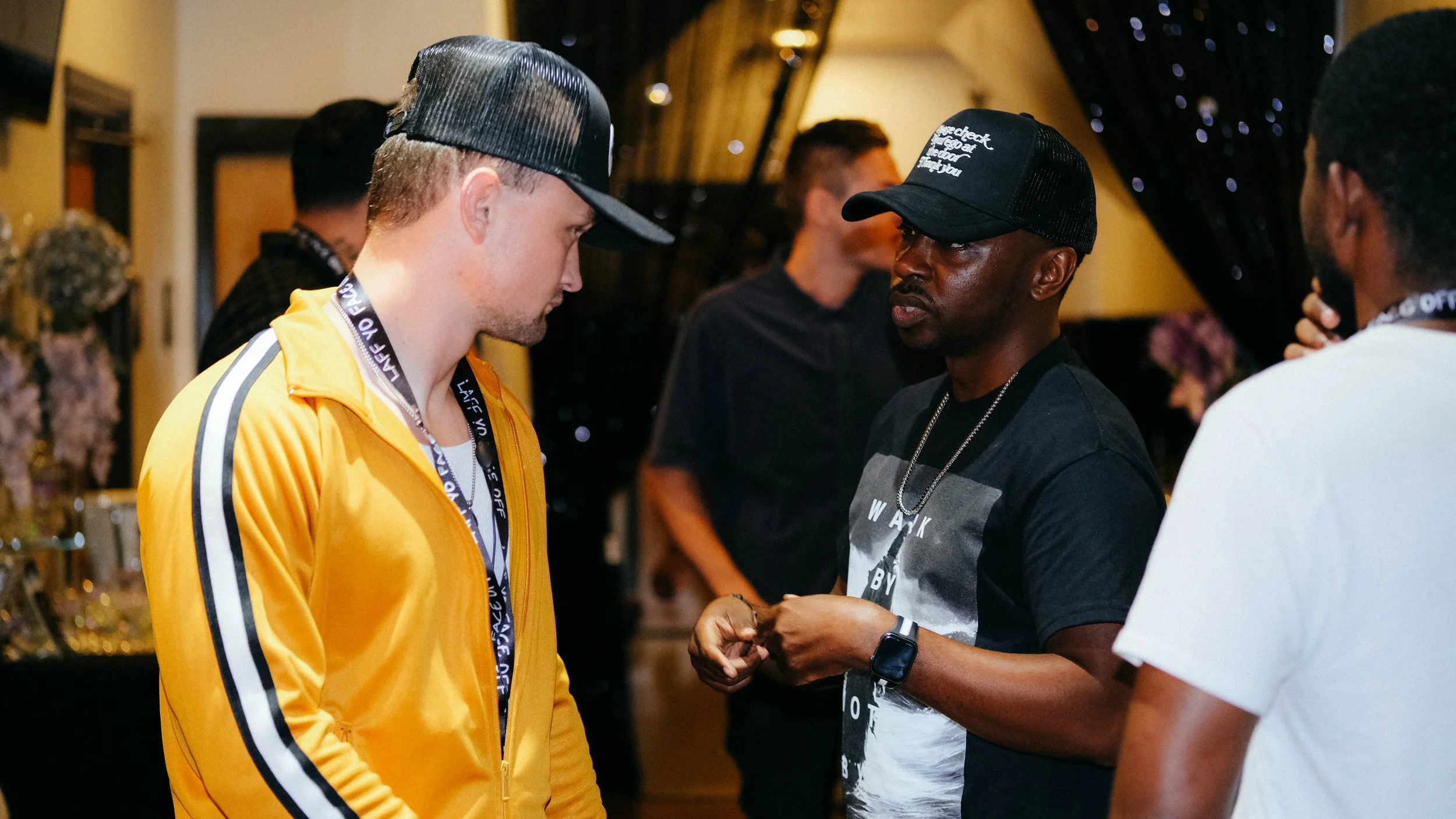 Two men are having a conversation in a social setting, with one wearing a yellow jacket and the other wearing a black cap. There are other people in the background, and the scene appears to be indoors with dim lighting.