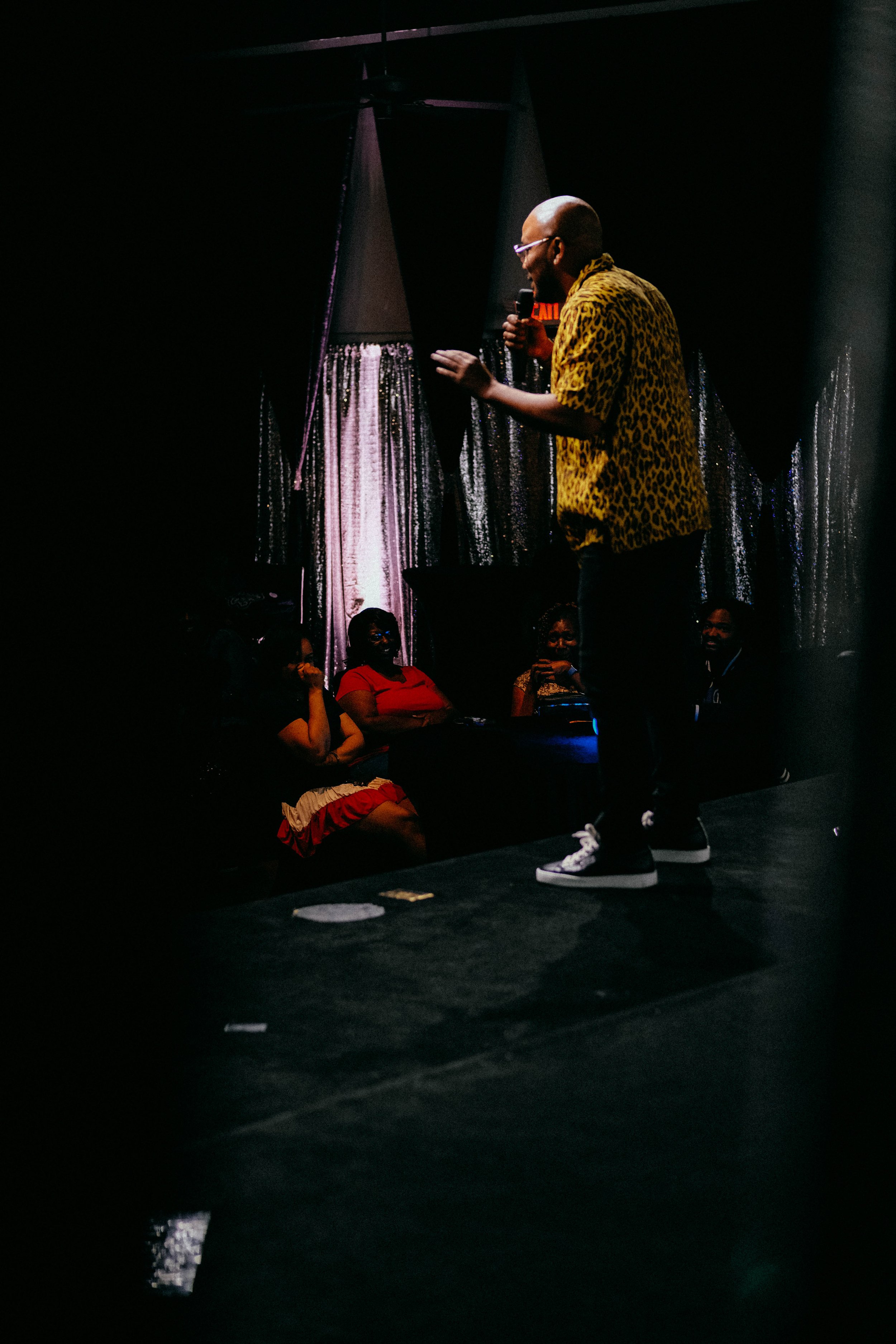 A man wearing a yellow leopard print shirt and white sneakers is speaking or singing into a microphone on stage at a dark venue, with a small audience seated in front of him.