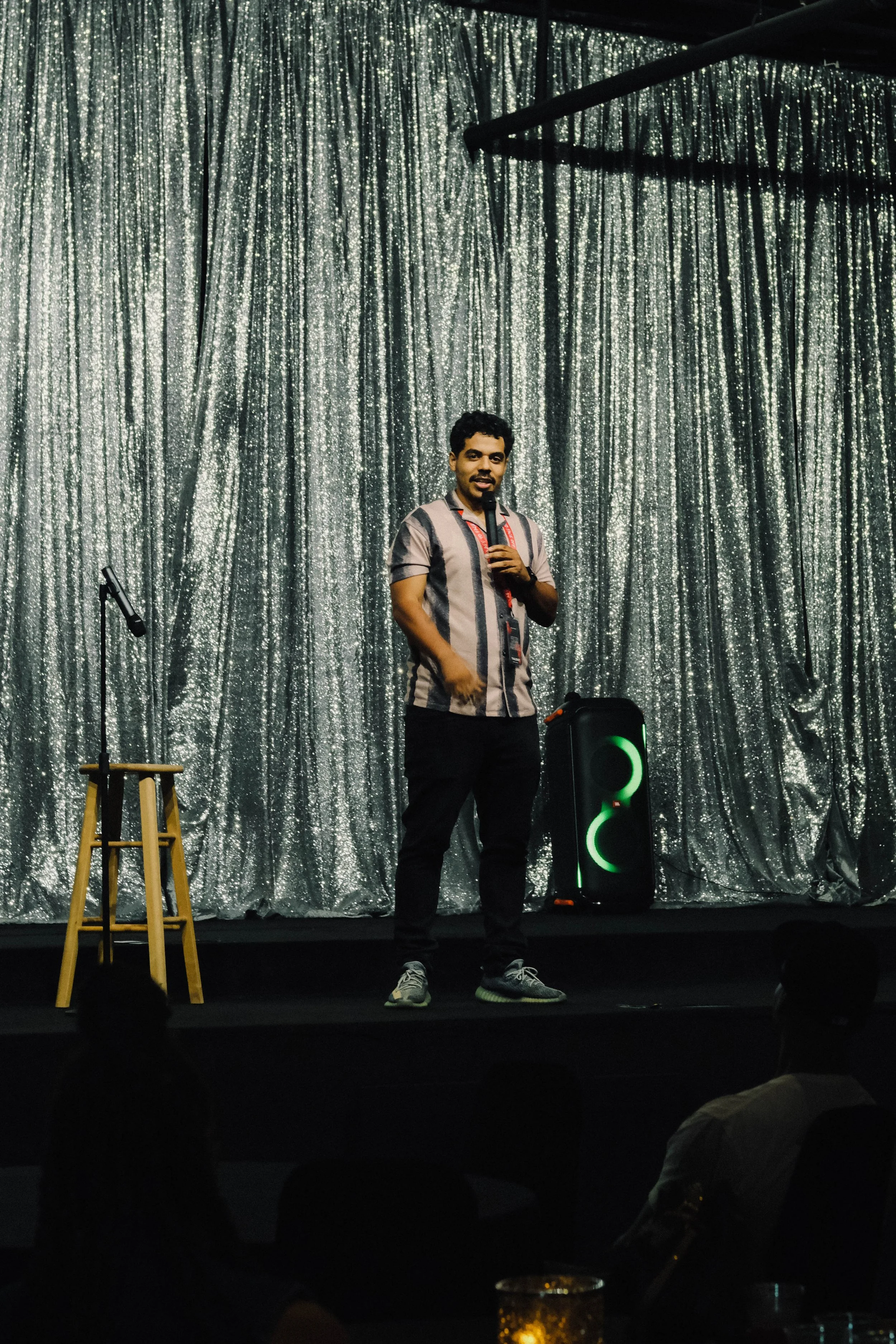 A man standing on a stage with a microphone in his hand, dressed in a striped shirt and black pants, in front of a silver, glittery curtain backdrop, with a stool and a speaker nearby, and audience silhouettes in the foreground.