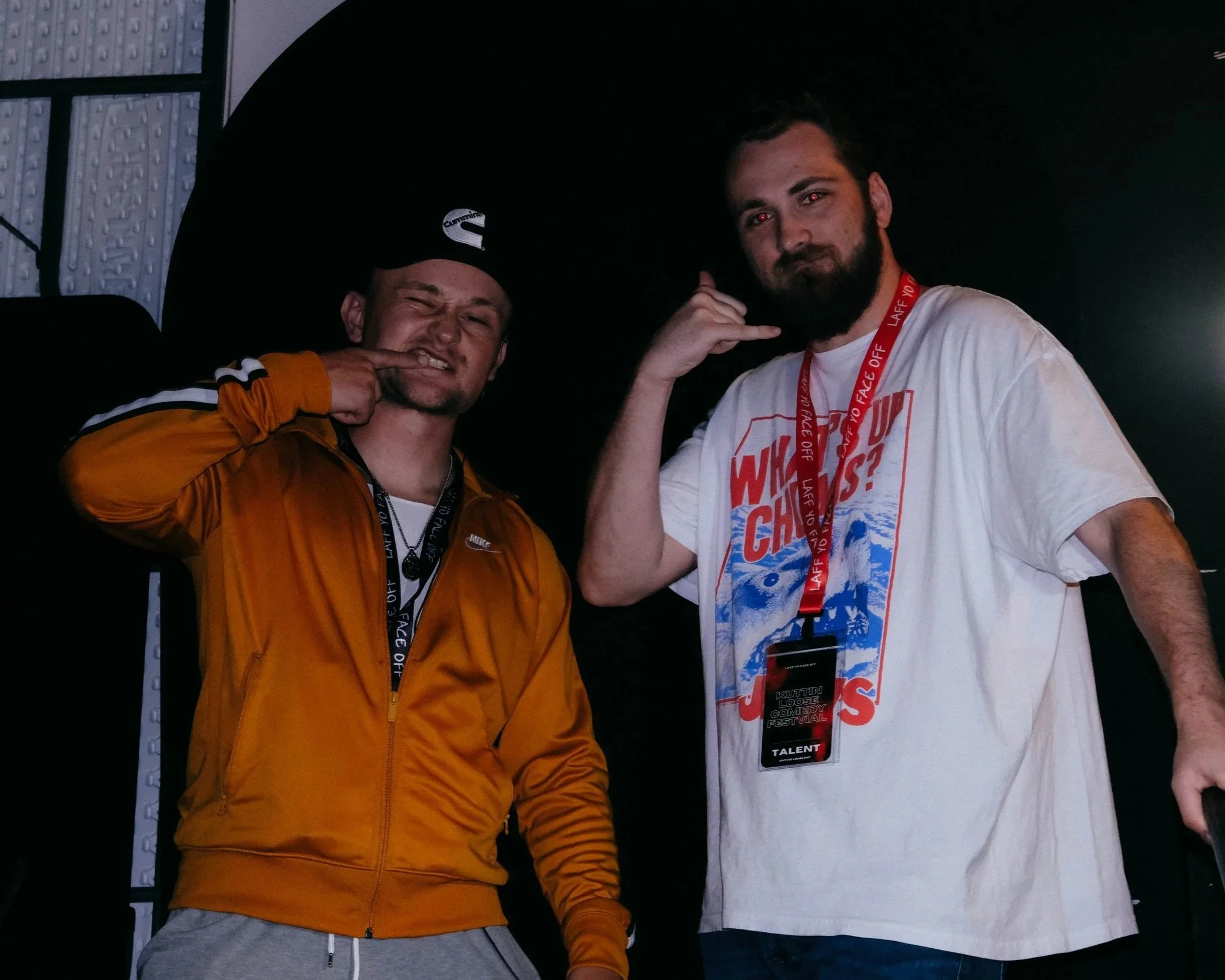 Two young men posing for a photo; the man on the left is wearing an orange jacket and gray pants, winking and pointing to his face; the man on the right is wearing a white T-shirt and a red lanyard, posing with his hand near his face.