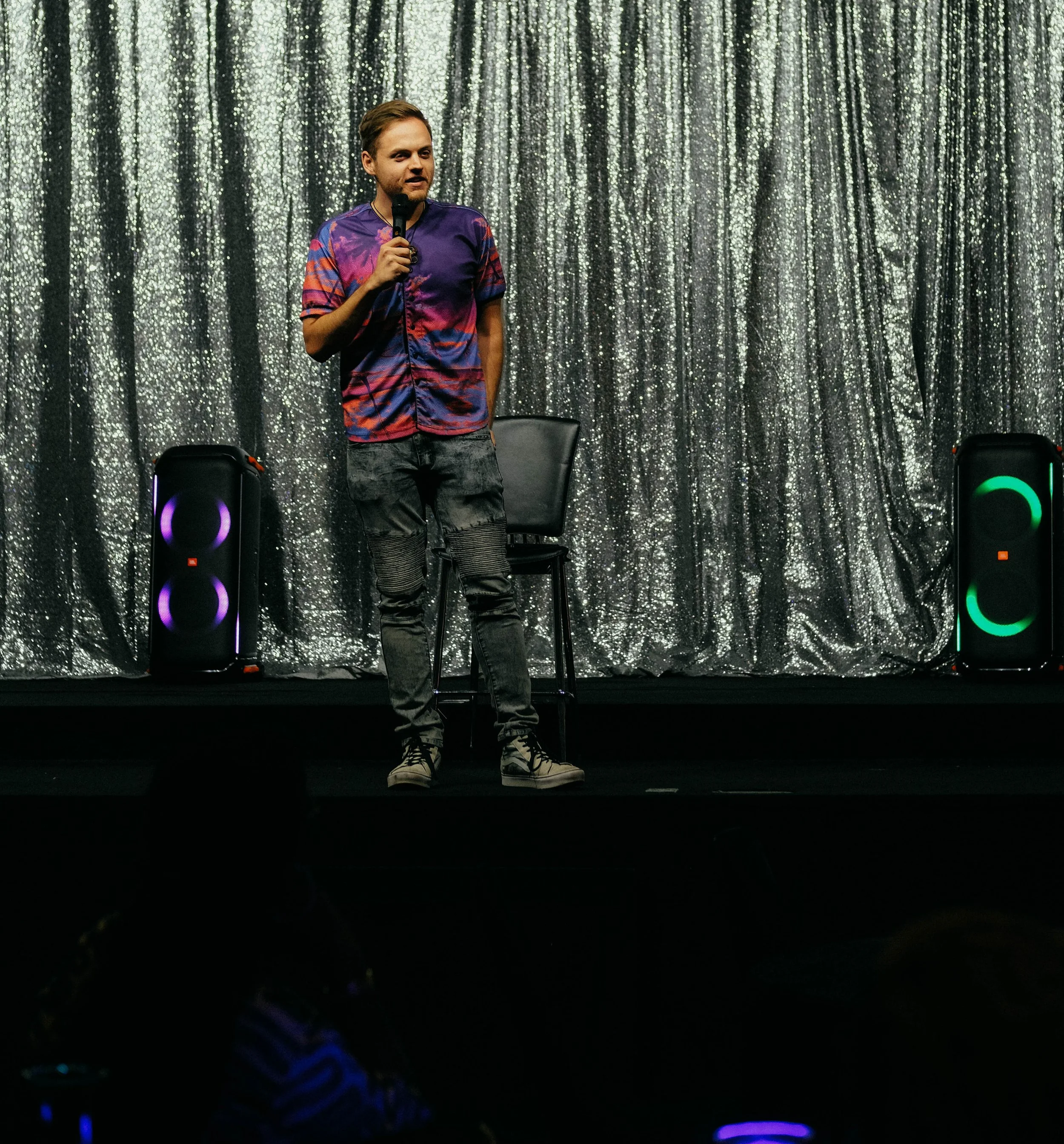 A man standing on a stage with a silver, glittery curtain background, holding a microphone and speaking, with two large black speakers on the stage.