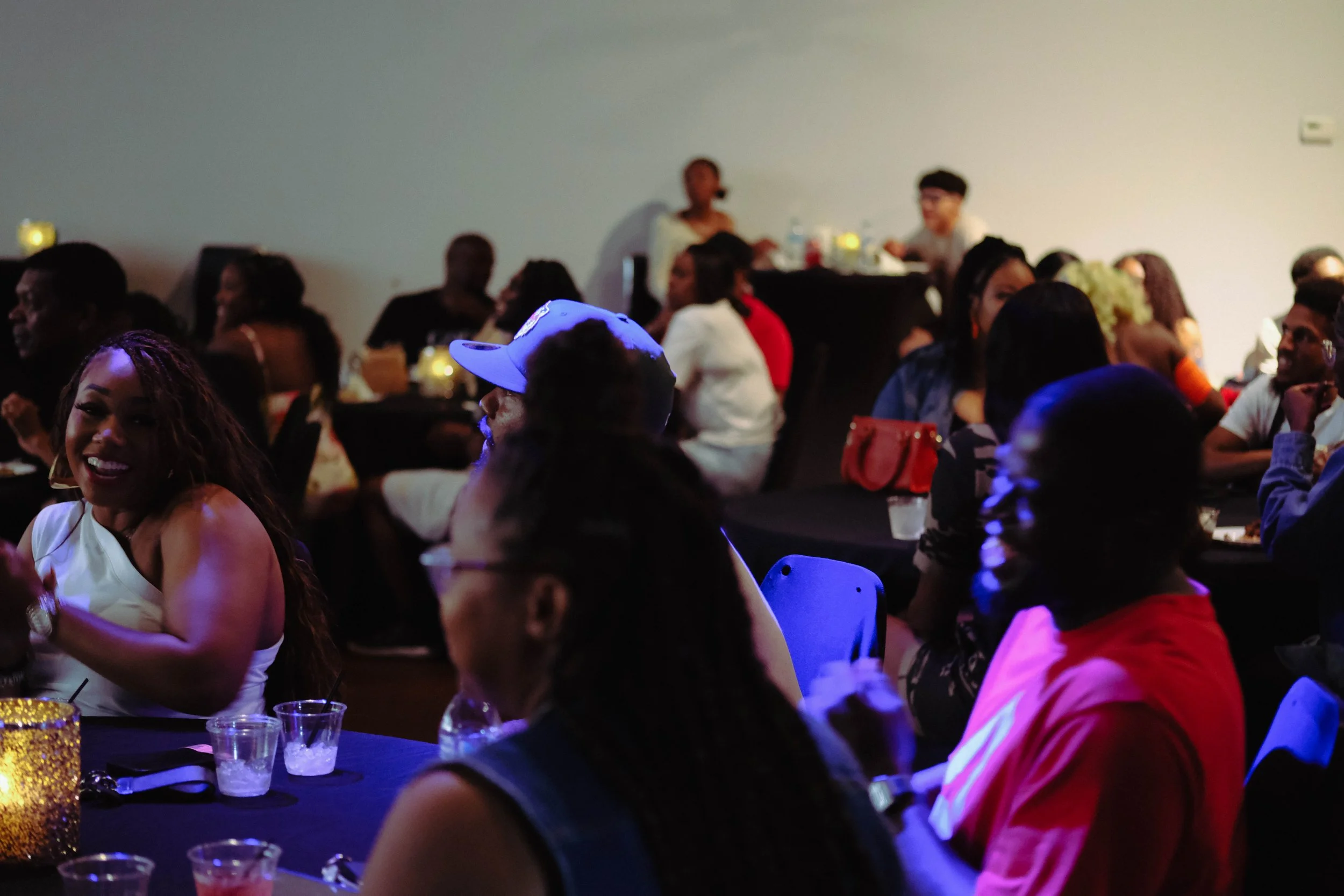 A crowded indoor party with people sitting at tables and socializing, some standing and talking. The scene includes a woman smiling in the foreground, and a woman standing at a bar in the background, with festive decorations and drinks on the tables.