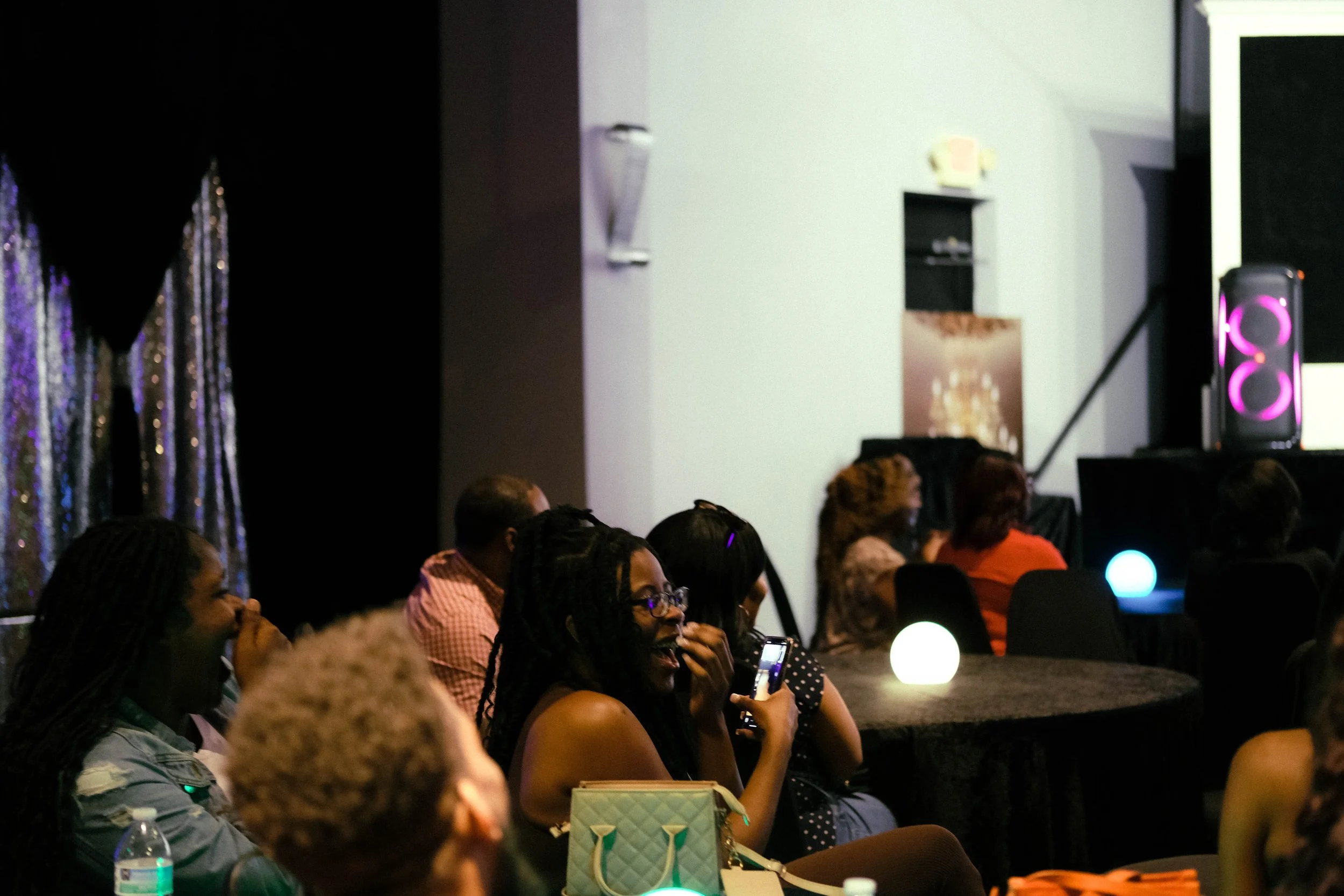 Audience seated at a dark room event, looking towards a stage or focal point outside the image frame, with some using their phones. There are water bottles on the tables, and the room has a black curtain with shiny, reflective strands on the left side and a speaker with pink lights on the right.