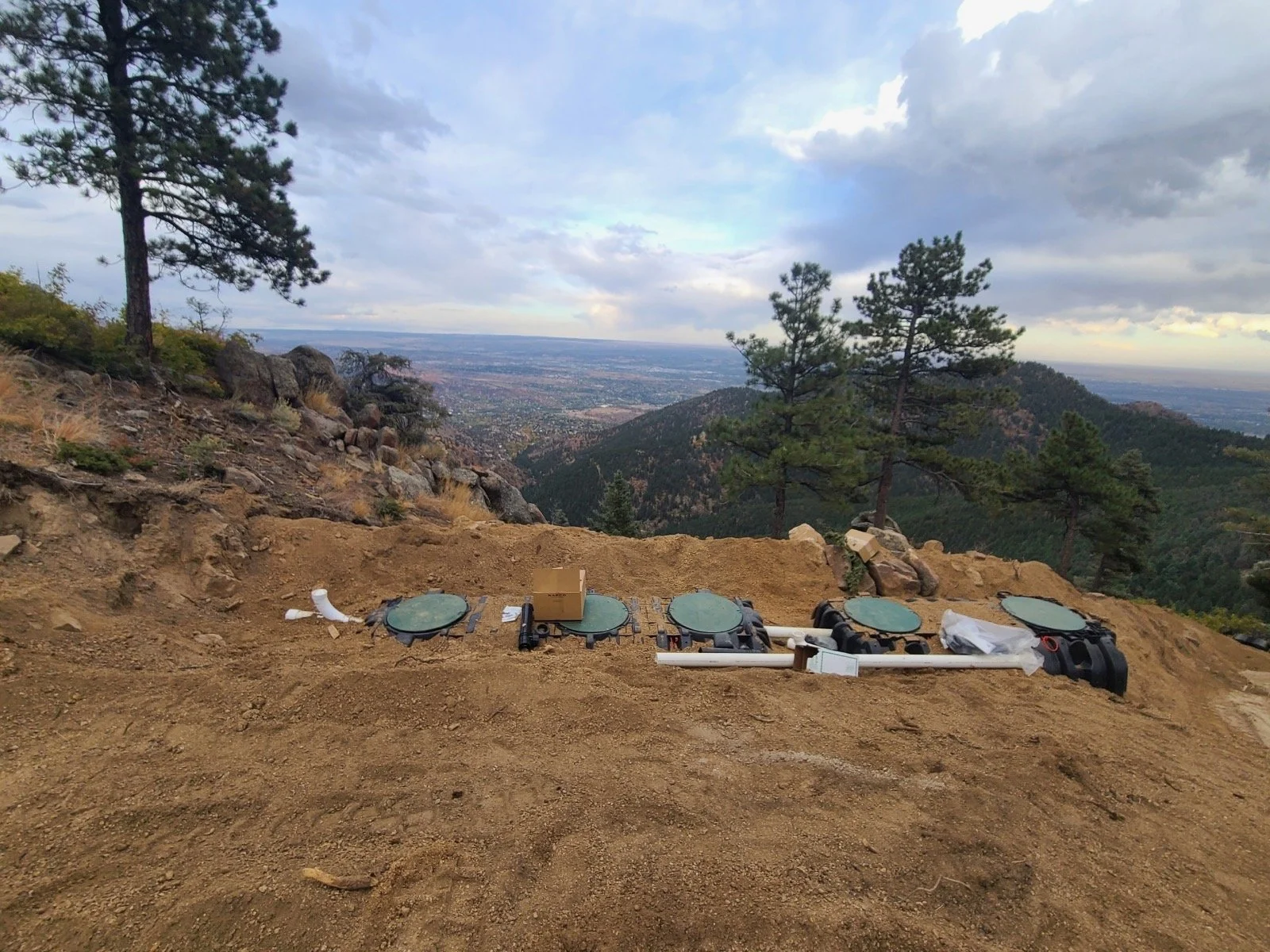 Construction site with plumbing pipes and fittings on dirt ground on a mountain, with a scenic view of trees, hills, and a cloudy sky in the background.